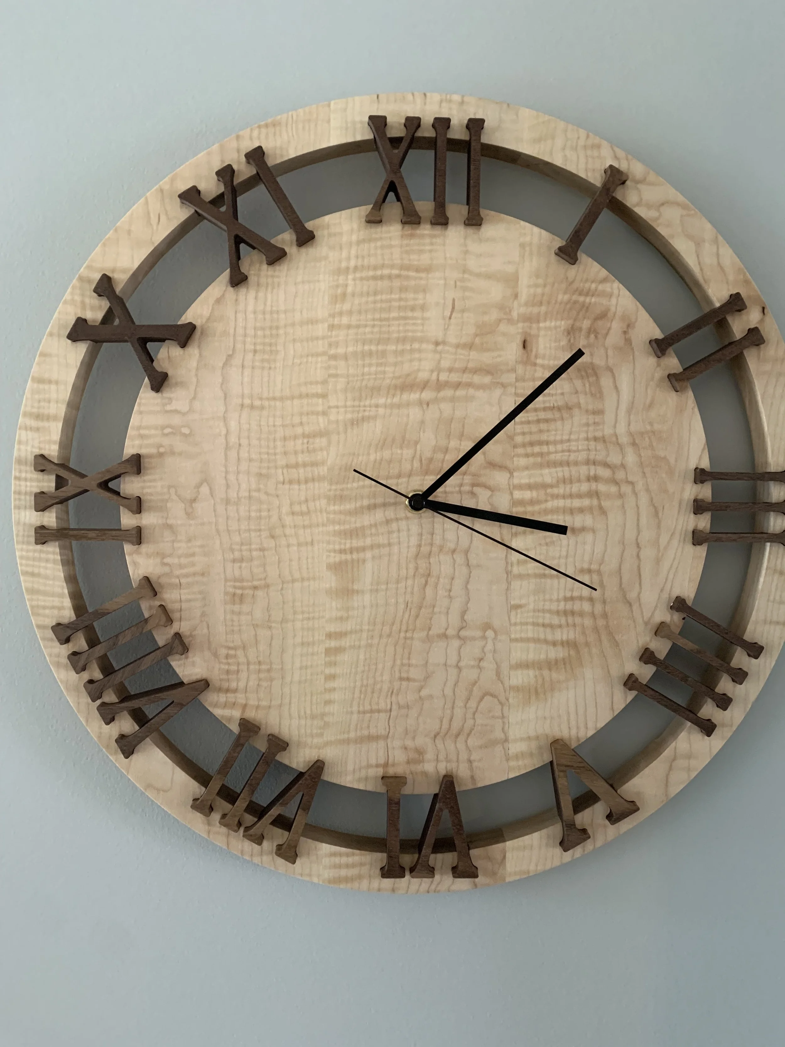 A round wooden wall clock with Roman numeral hour markers made of dark wood on a light wood face. The clock shows the time as approximately 3:24 with two black hands and a thin black second hand.