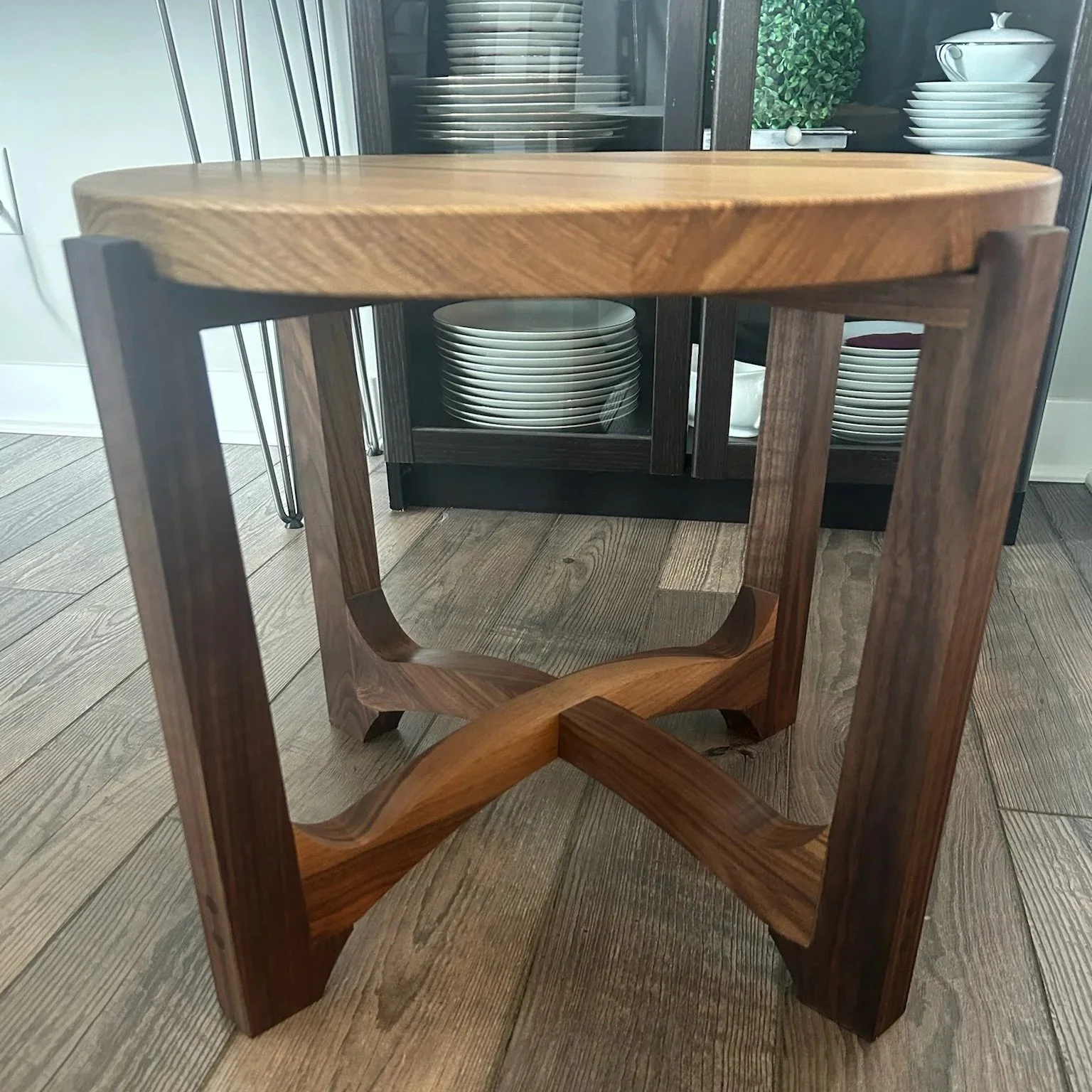 A wooden stool with a curved base, viewed from above in front of a dark cabinet filled with plates and bowls.