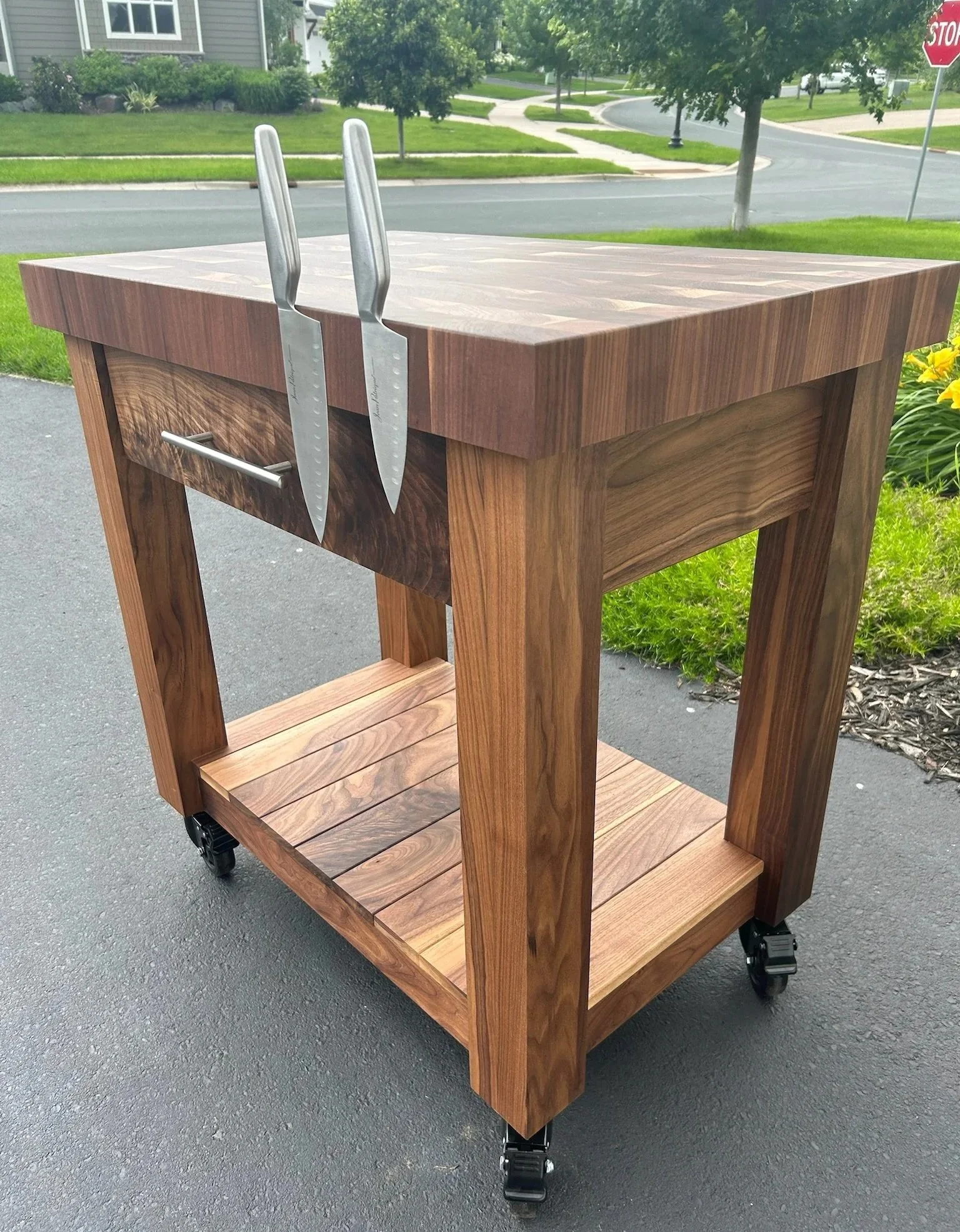 A wooden rolling cart with a butcher block top, in an outdoor setting on a paved surface, with a background of grass, trees, houses, and a stop sign.