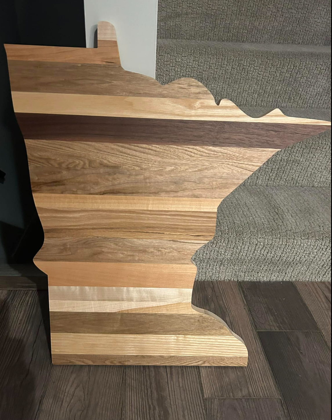 Wooden Minnesota-shaped decor piece with a natural wood finish, placed on a wooden floor next to a textured beige carpeted staircase.