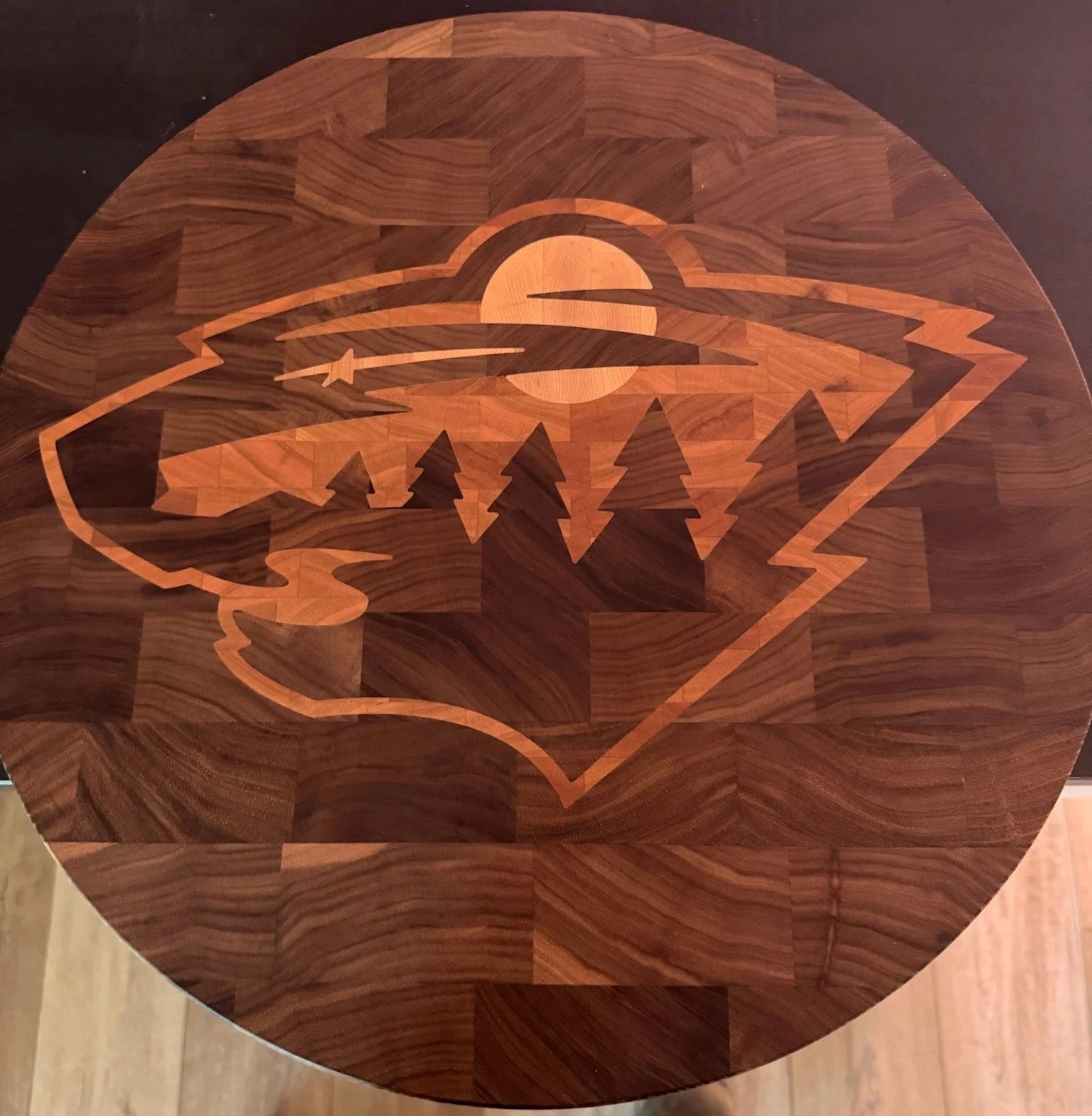 A round wooden table with an inlaid design of a wolf's head, howling at a rising or setting sun, with a background of trees and clouds.