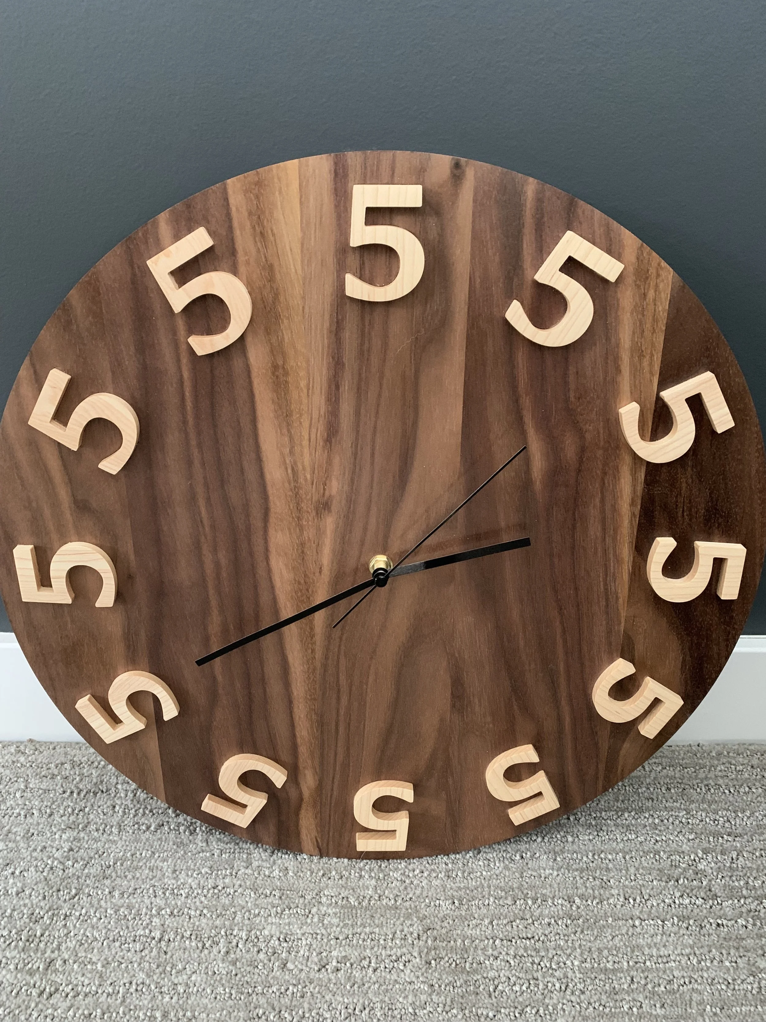 A round wooden clock with beige 3D numbers on a gray wall, sitting on a carpeted floor.