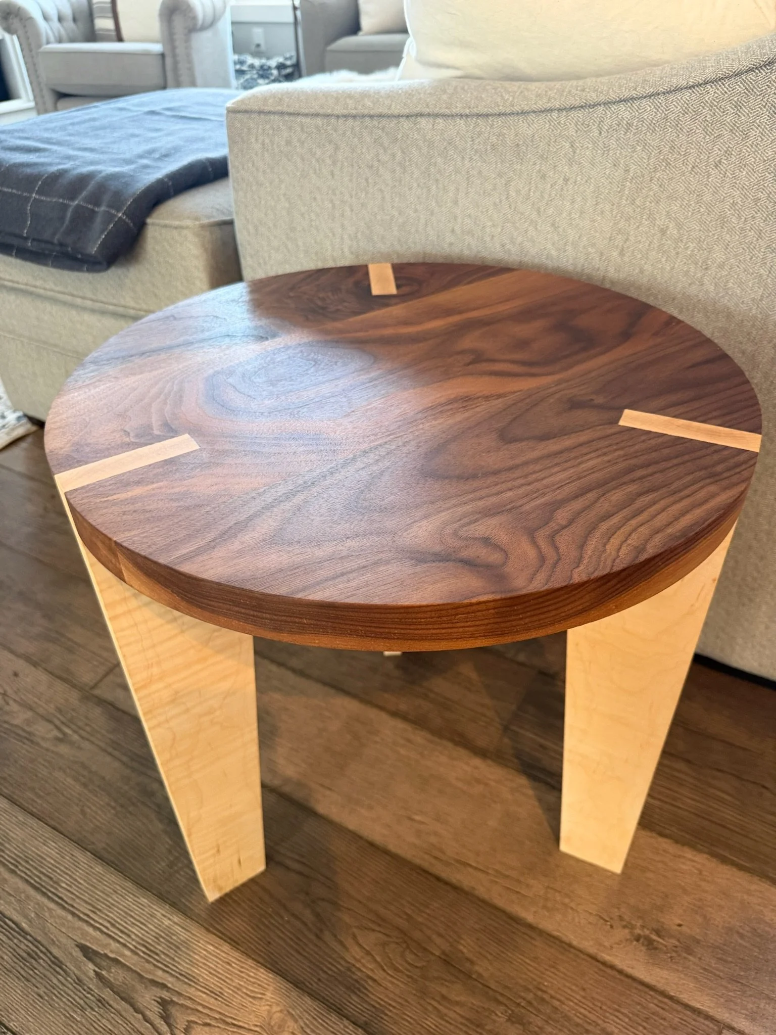 A wooden side table with a dark wood top and light wood legs, placed next to a beige upholstered armchair in a living room setting.