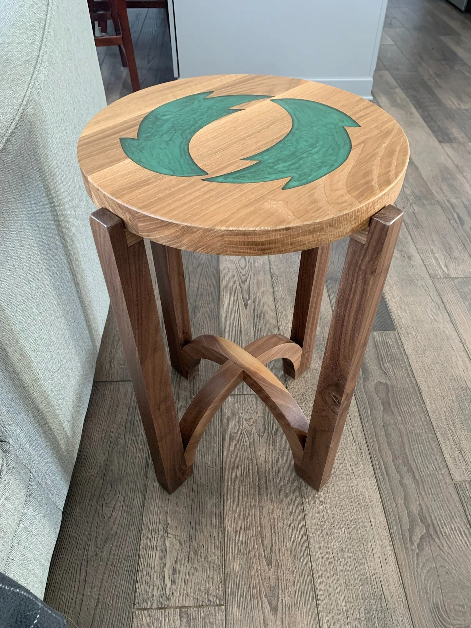 Round wooden side table with a green circular design on top, next to a beige sofa and a brown wooden chair in a room with wood flooring.