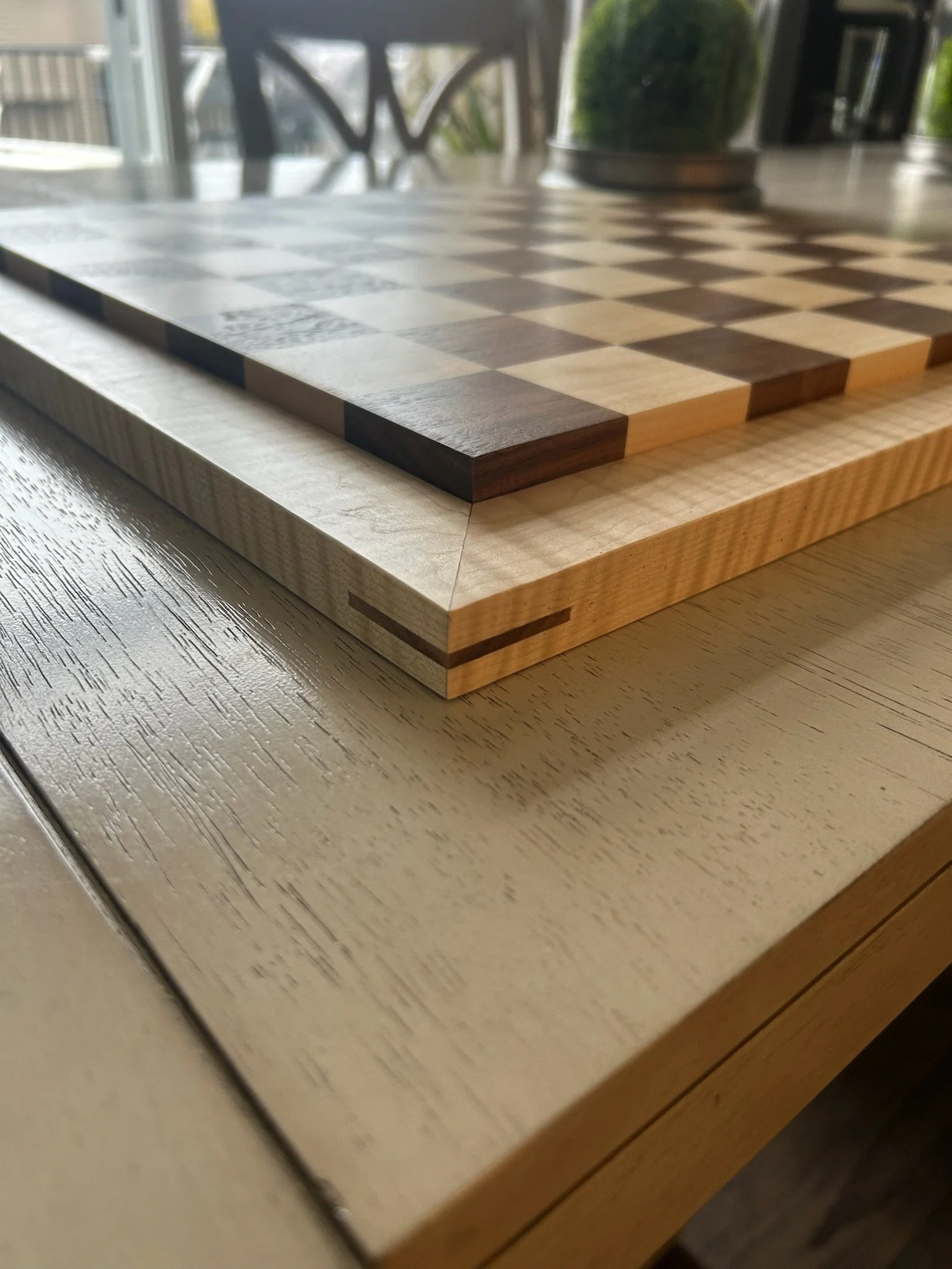 Close-up view of a wooden chessboard on a table indoors, with a blurred background of a window, chairs, and houseplants.
