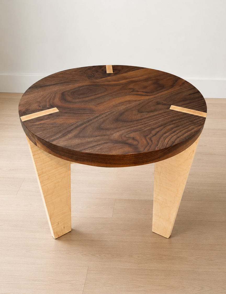 Wooden side table with a dark round top and light angled legs, placed on light wood flooring against a white wall.