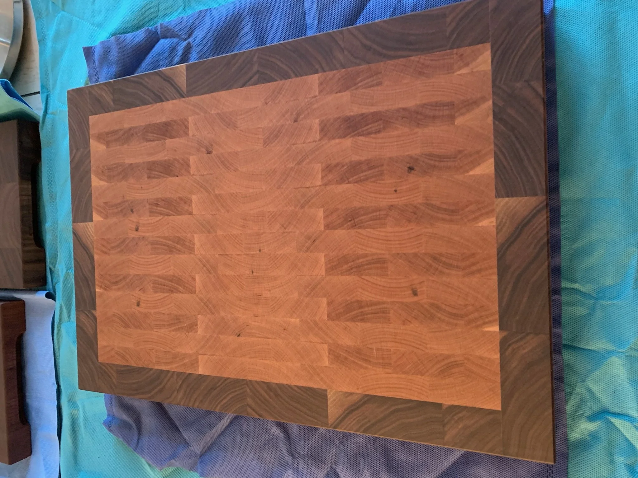 A square wooden table with a pattern of light and dark wood grains, placed on a bed with a blue and green blanket.