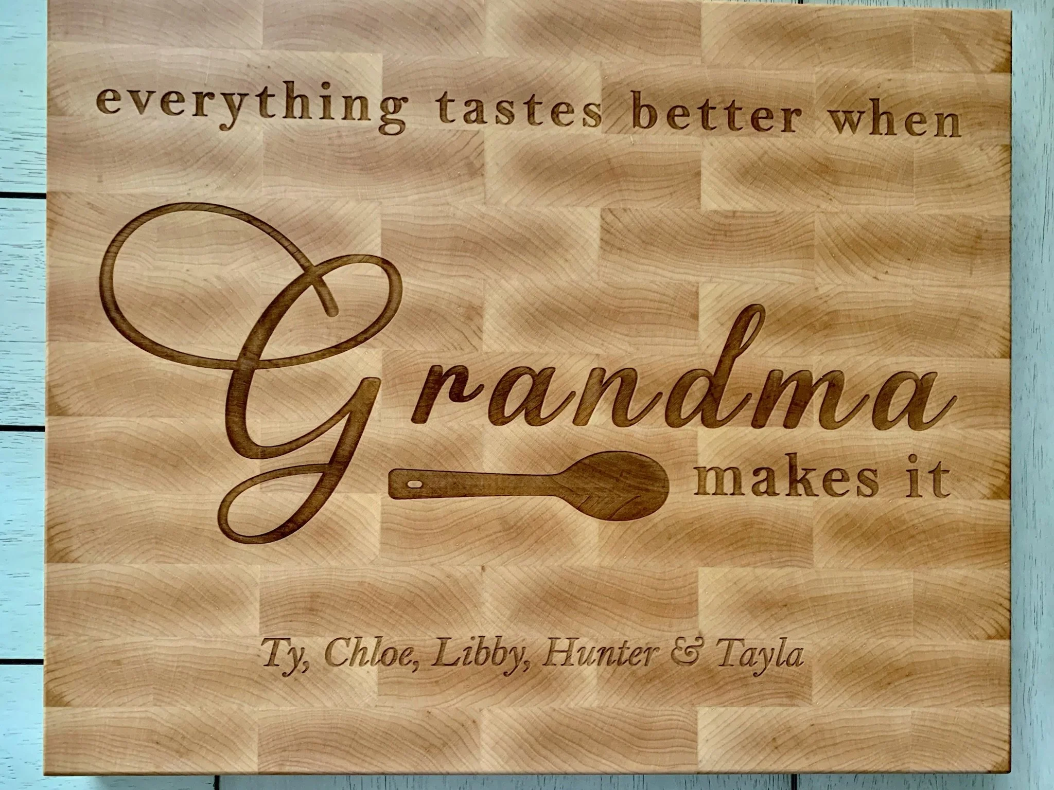 A wooden plaque with engraved text reading 'everything tastes better when grandma makes it' and a small spoon illustration, placed on a turquoise table with a slatted design.