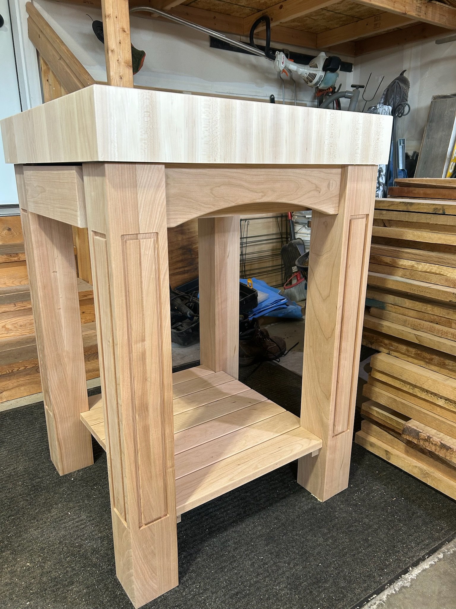 A wooden furniture piece in progress, likely a table or stand, with a square top and sturdy legs, situated in a workshop with tools and lumber around.