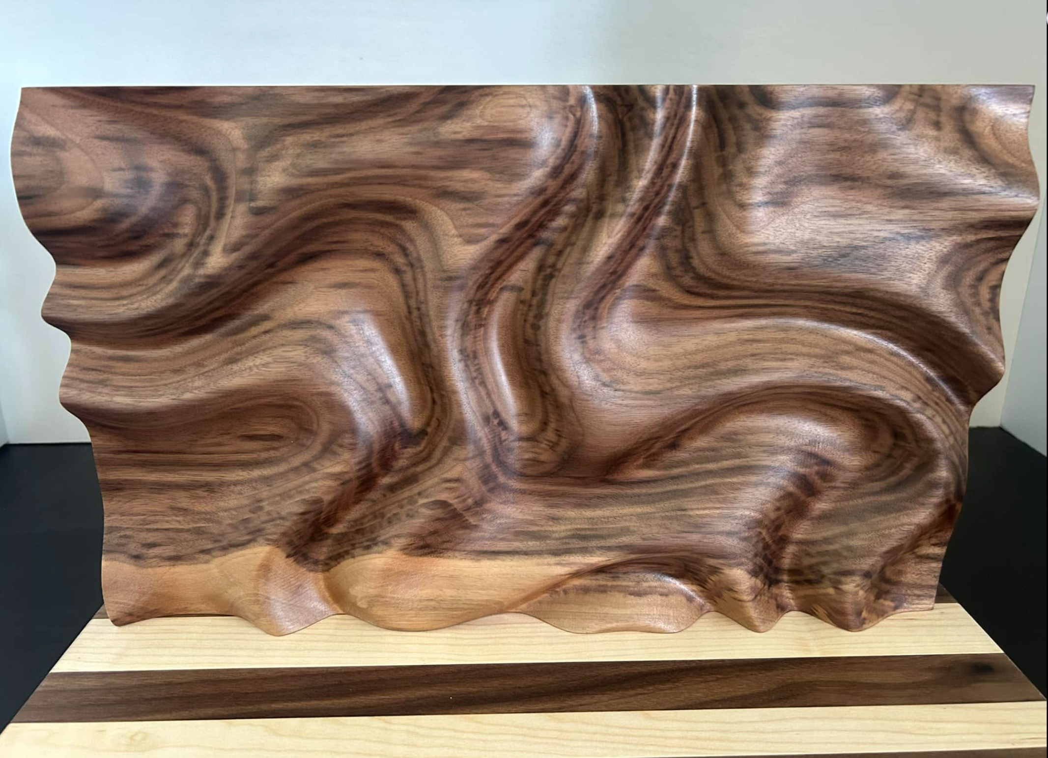 A wooden sculpture with wavy, flowing grain patterns, placed on a light wooden base.