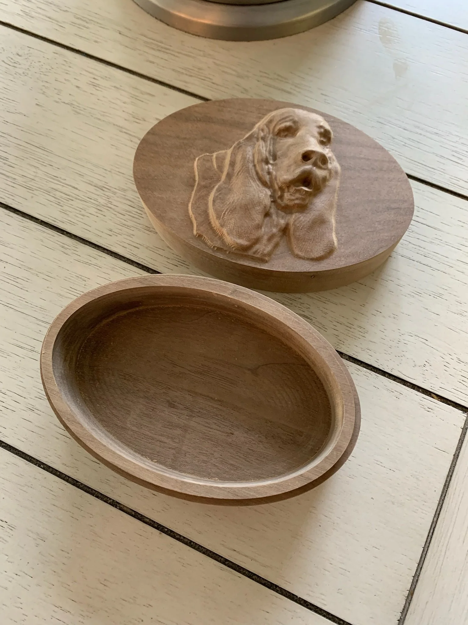 A wooden box with a carved image of a dog's head on its lid, and an empty wooden bowl, placed on a light-colored wooden table.