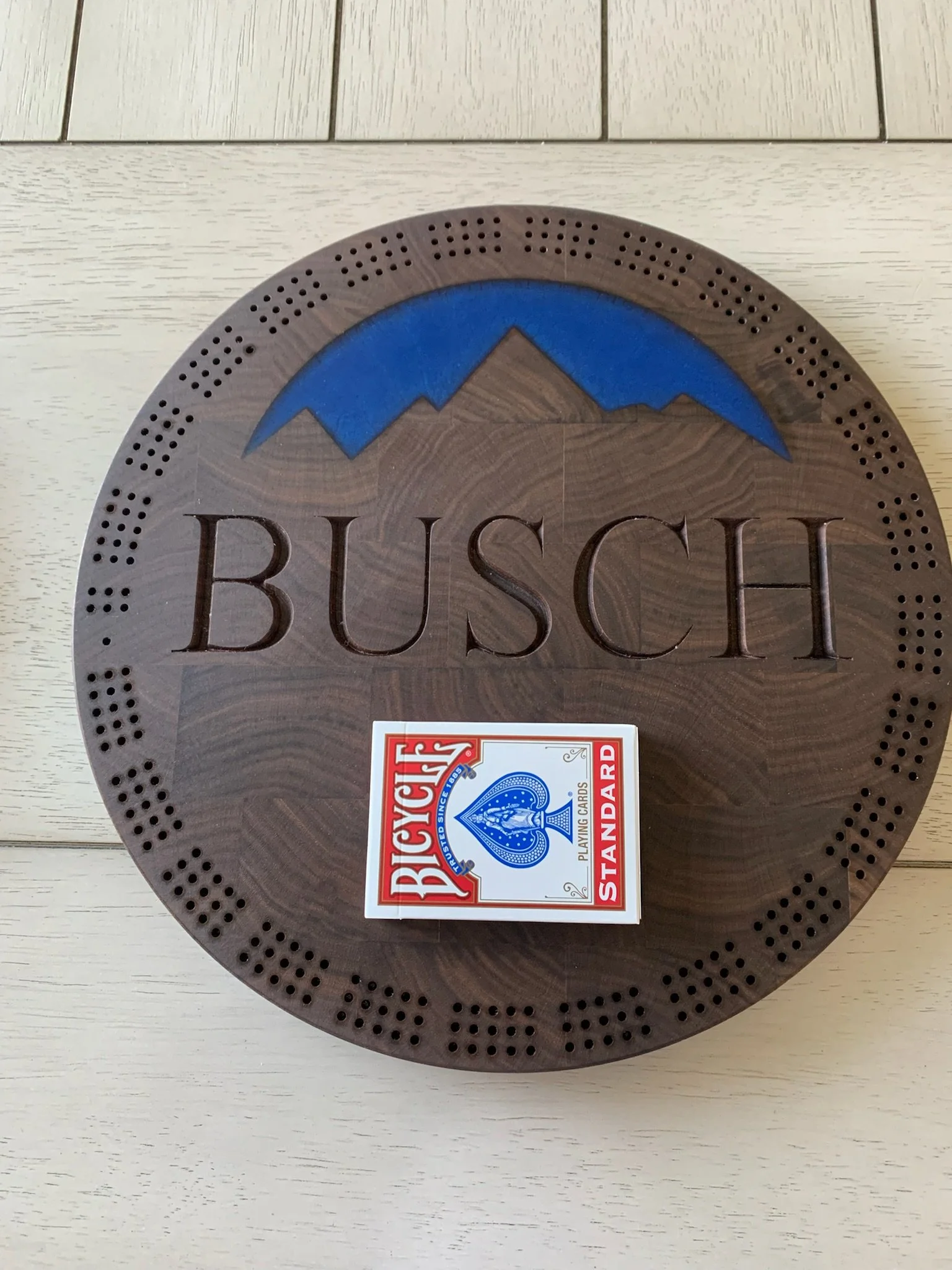 Round wooden plaque with a mountain and blue sky design at the top, the word 'BUSH' engraved in large letters, and a deck of Bicycle standard playing cards placed on it.