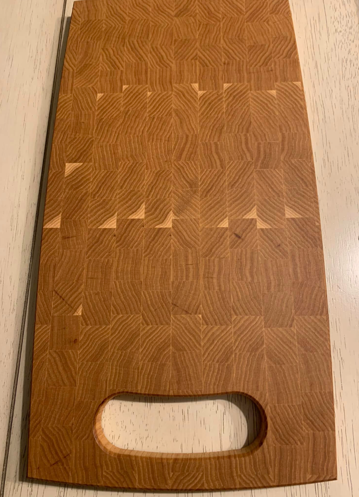 A wooden cutting board with a rectangular shape, rounded edges, and a handle with a hole at the bottom, resting on a white wooden surface.
