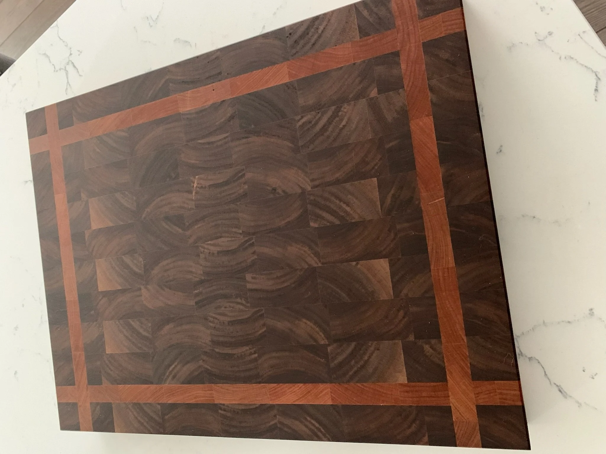 The image shows a close-up of an intricate wooden floor with a decorative pattern composed of dark and reddish-brown wood planks arranged in a square mosaic with a border, beside a white marble surface with gray veining.