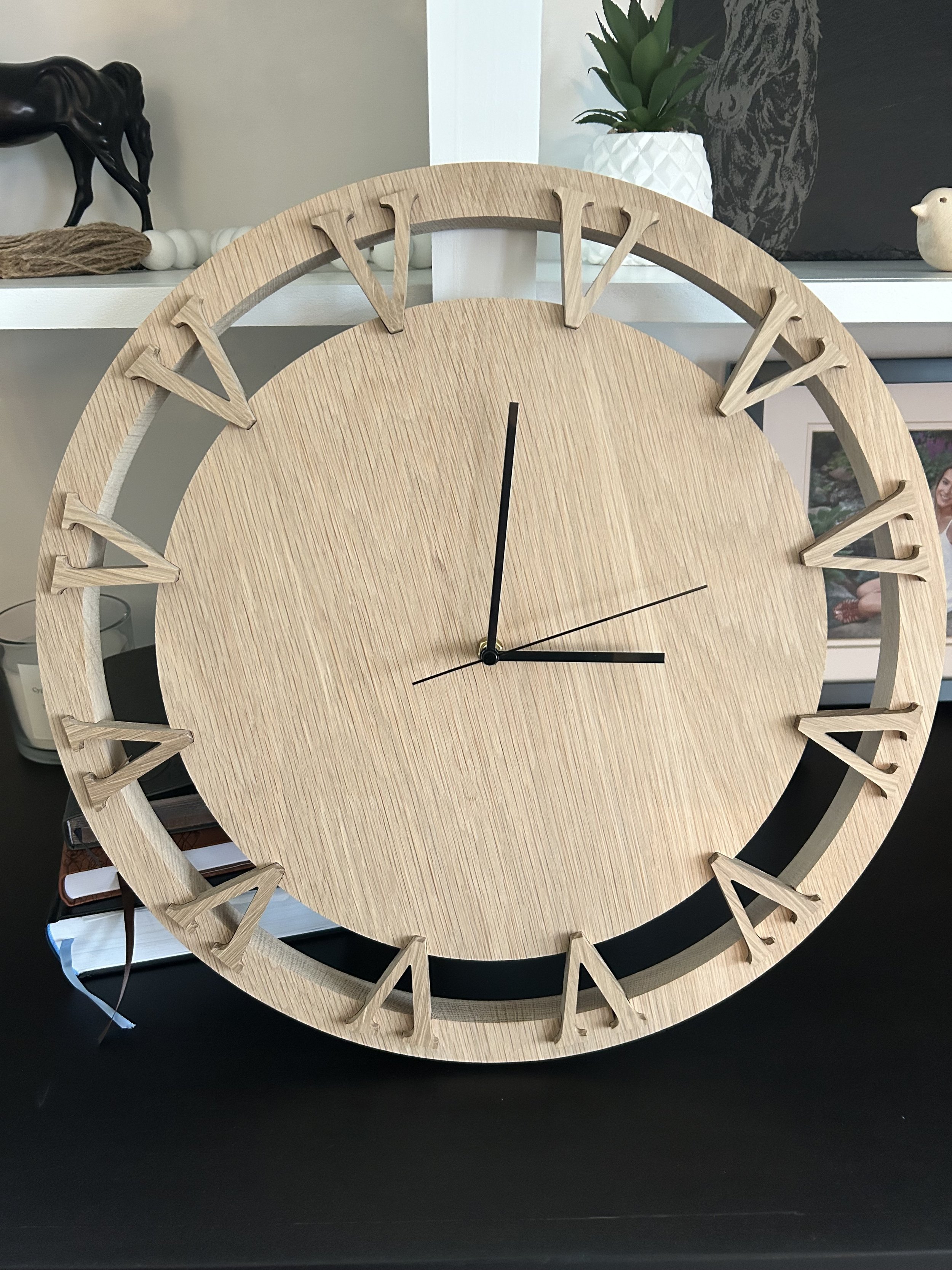 Close-up of a round wooden wall clock with Roman numerals, black hour and minute hands, and a black second hand, placed on a black surface. In the background, there are decorative items including a potted plant, a small bird figurine, and framed phot