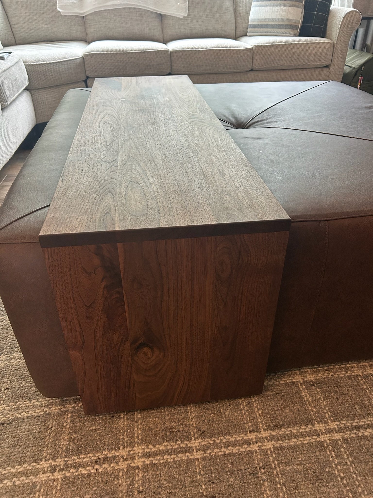 A large wooden desk with a textured top and detailed wood grain pattern situated in front of a beige upholstered sofa with cushions, on a brown carpet.