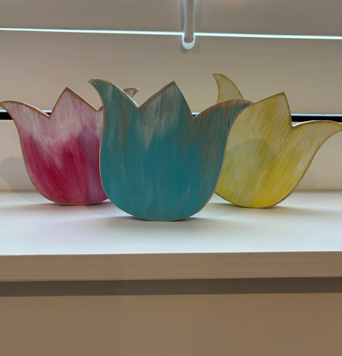 Three colorful wooden tulip-shaped decorative items, pink, blue, and yellow, placed on a white surface with a background of horizontal blinds.