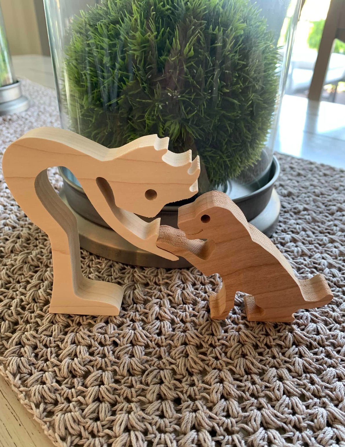 Wooden dinosaur figurines placed on a woven table runner in front of a potted green plant with small leaves.