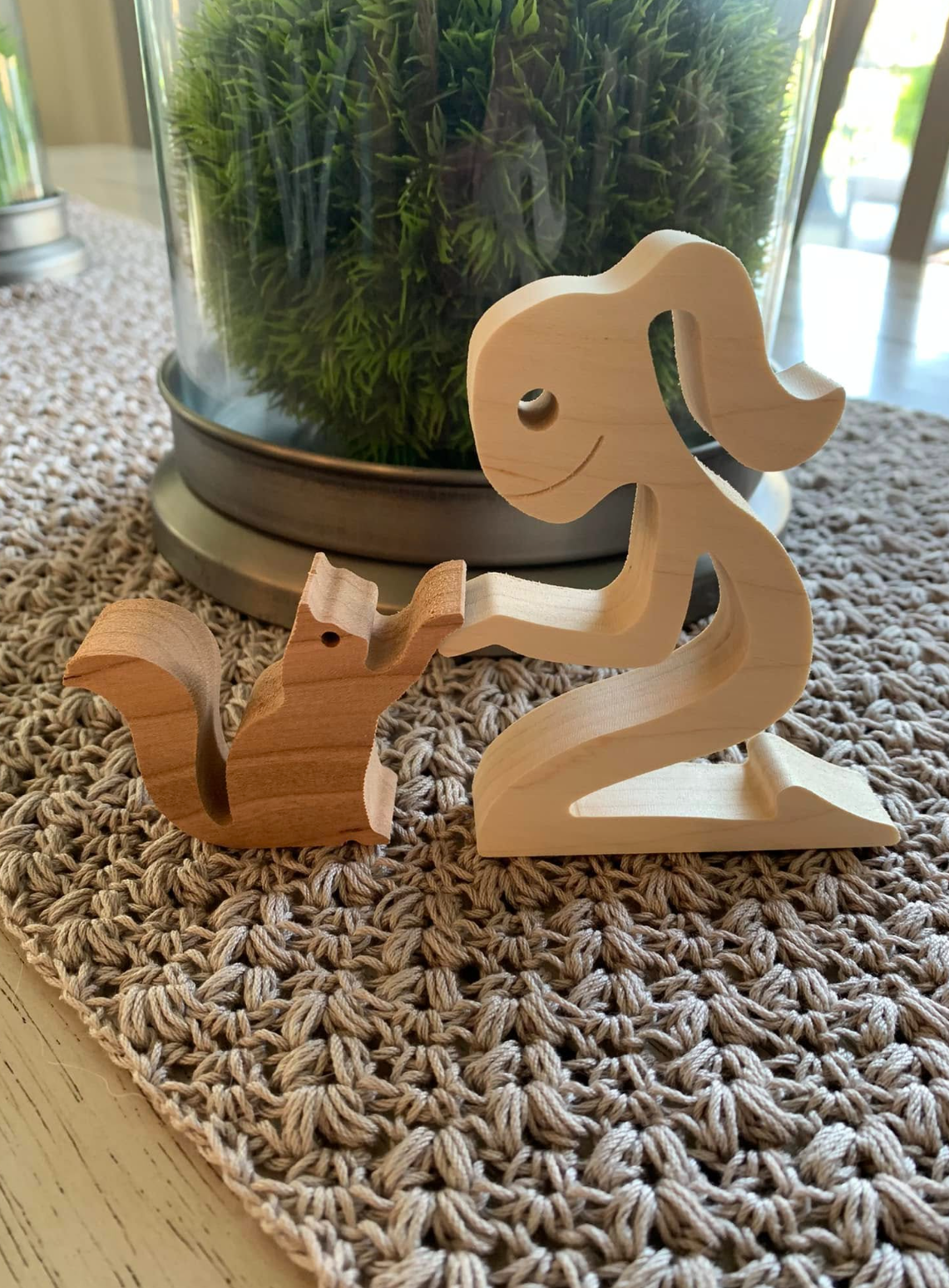 A wooden elephant-shaped number two decoration with a small wooden curly creature, placed on a woven beige table runner. In the background, there is a glass container with green plants and a metal base.