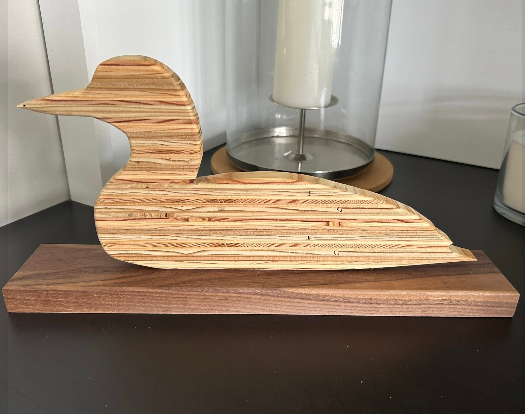 Wooden duck sculpture on a wooden base, with candles and a metal and glass lamp in the background.