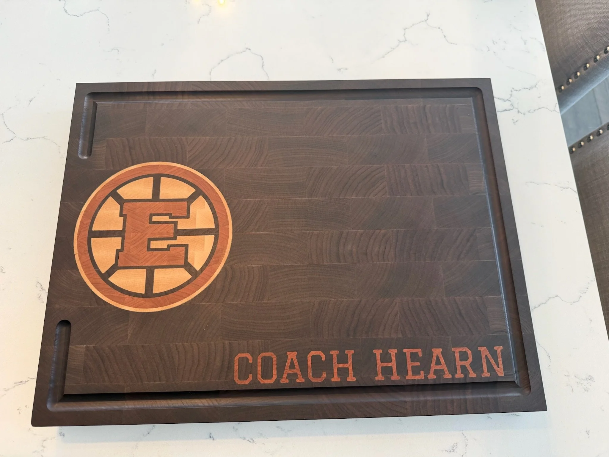 A wooden desk or serving tray with a Boston Bruins logo design, which features a black 'B' with yellow outline and a circle around it, placed on a white marble or quartz countertop with gray veining.