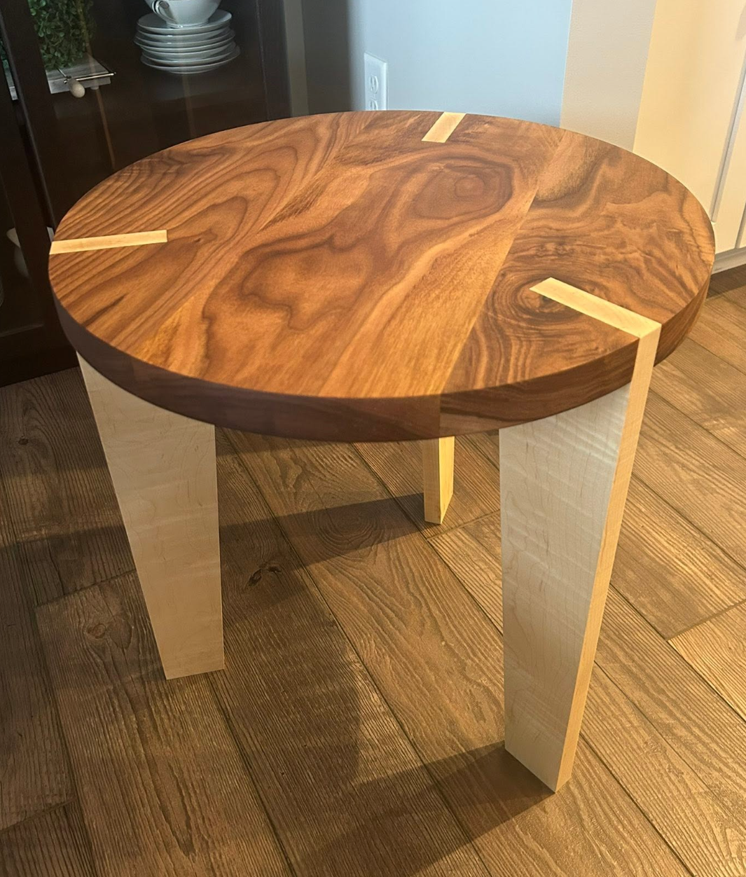 Round wooden table with a two-tone wood top and light-colored legs, placed on a wooden floor in a corner of a room.