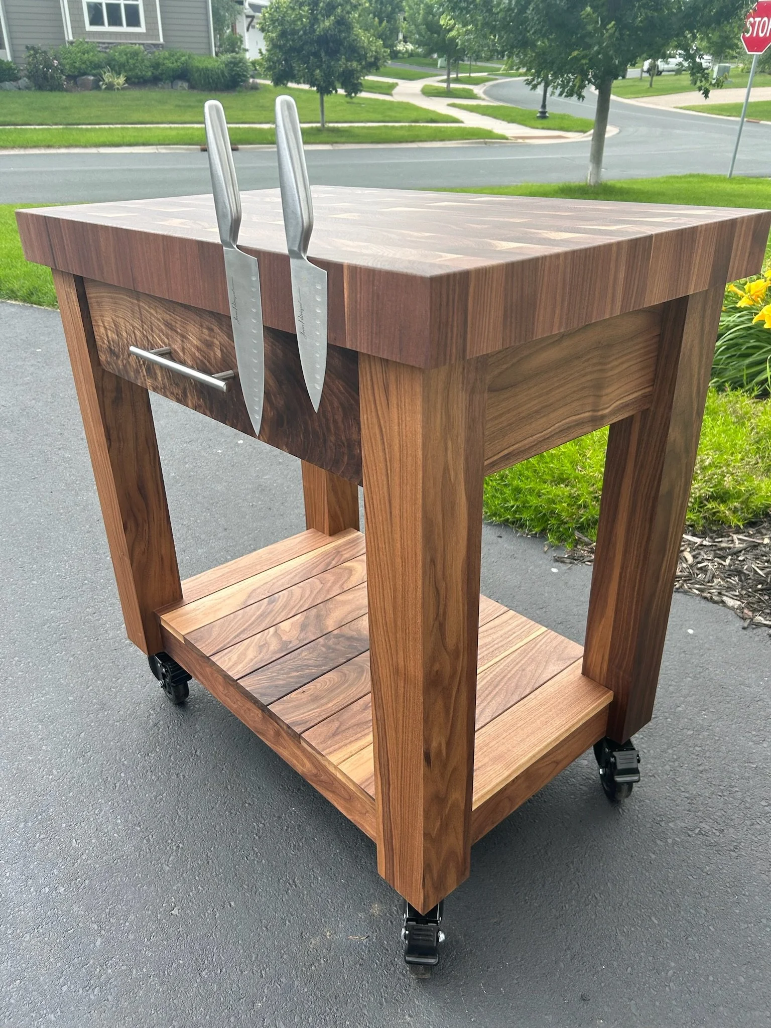 A wooden serving or kitchen cart on wheels with a butcherblock top, slatted shelf at the bottom, a handle on the side, and two knives hanging from the side.
