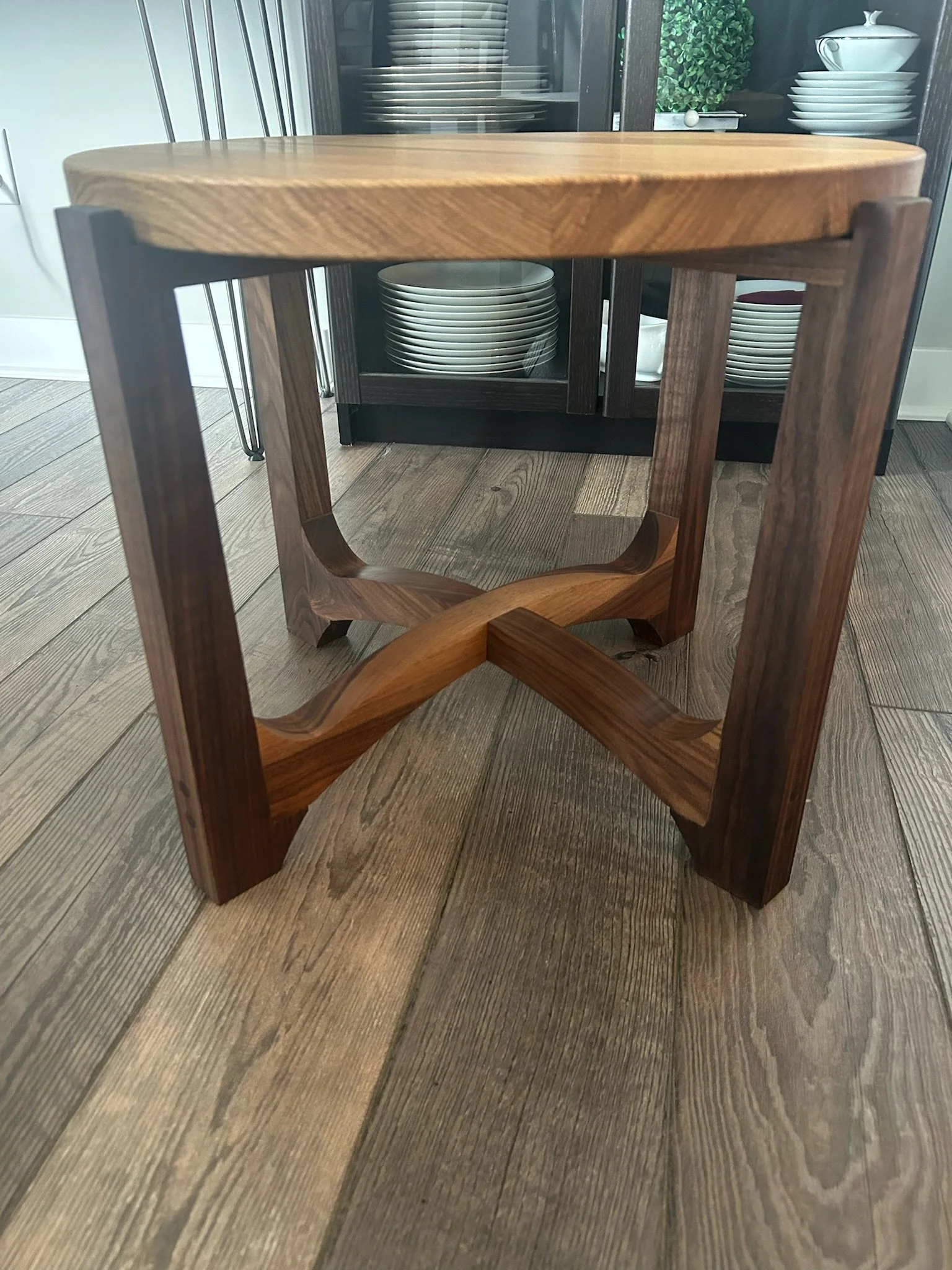 A close-up view of a wooden stool with crossed legs on a wooden floor, with a dark cabinet in the background holding dishes and a plant.