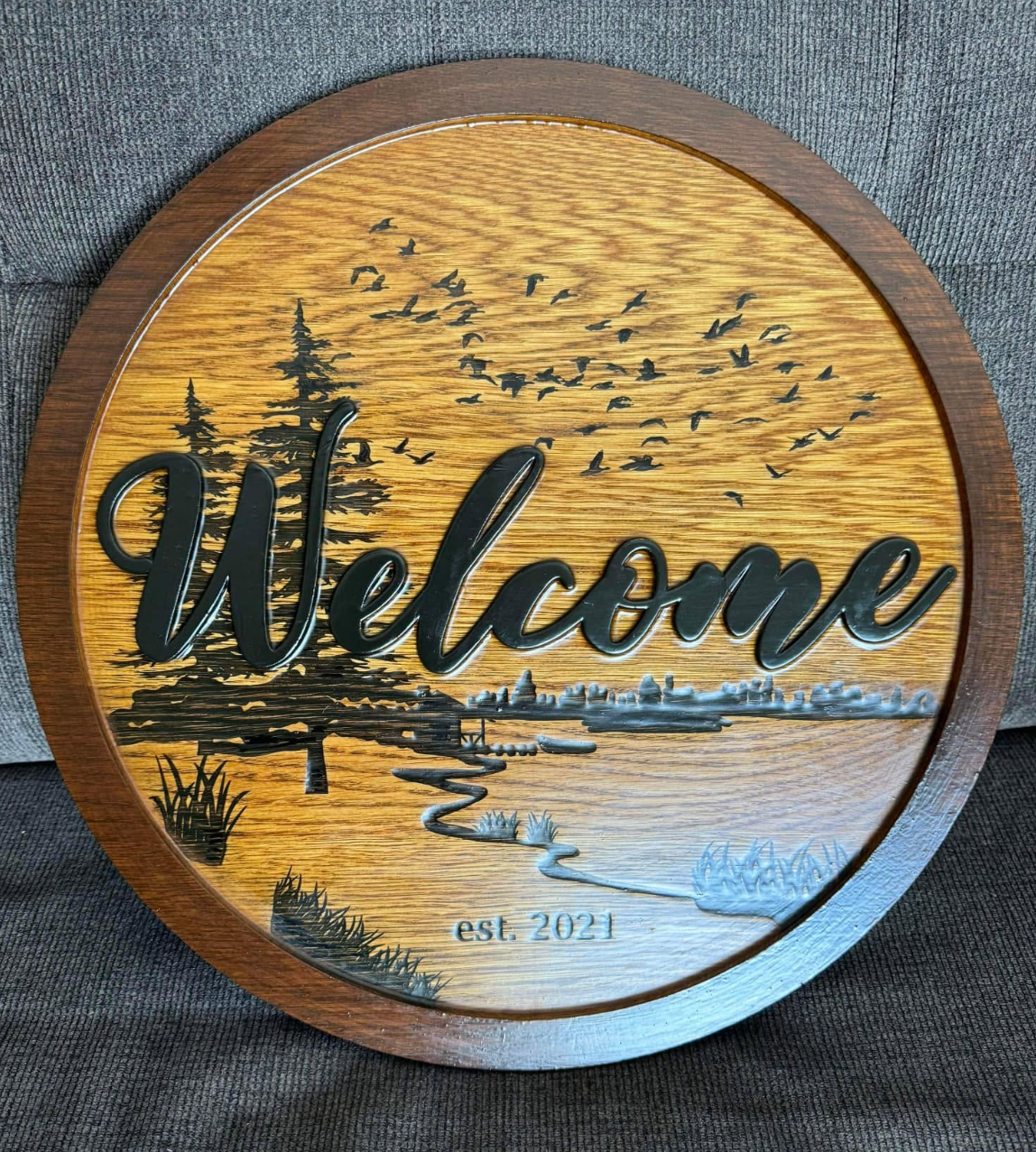 Decorative wooden wall hanging with the word 'Welcome' in large script, featuring a landscape with trees, a river, birds, and city in the background, and 'est. 2021' at the bottom.