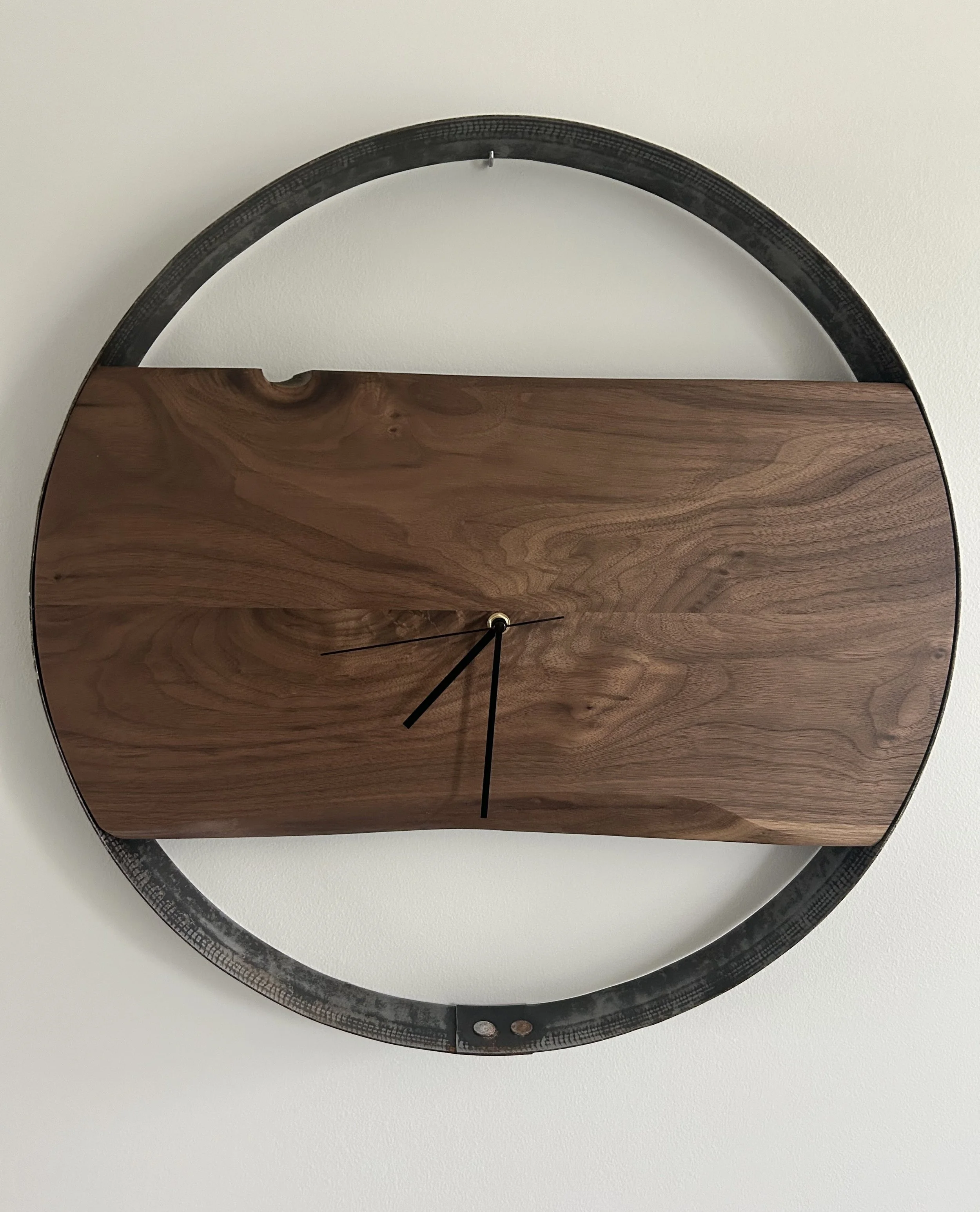 Wall clock with a square wooden face and black clock hands, mounted inside a black metal circular frame attached to a white wall.