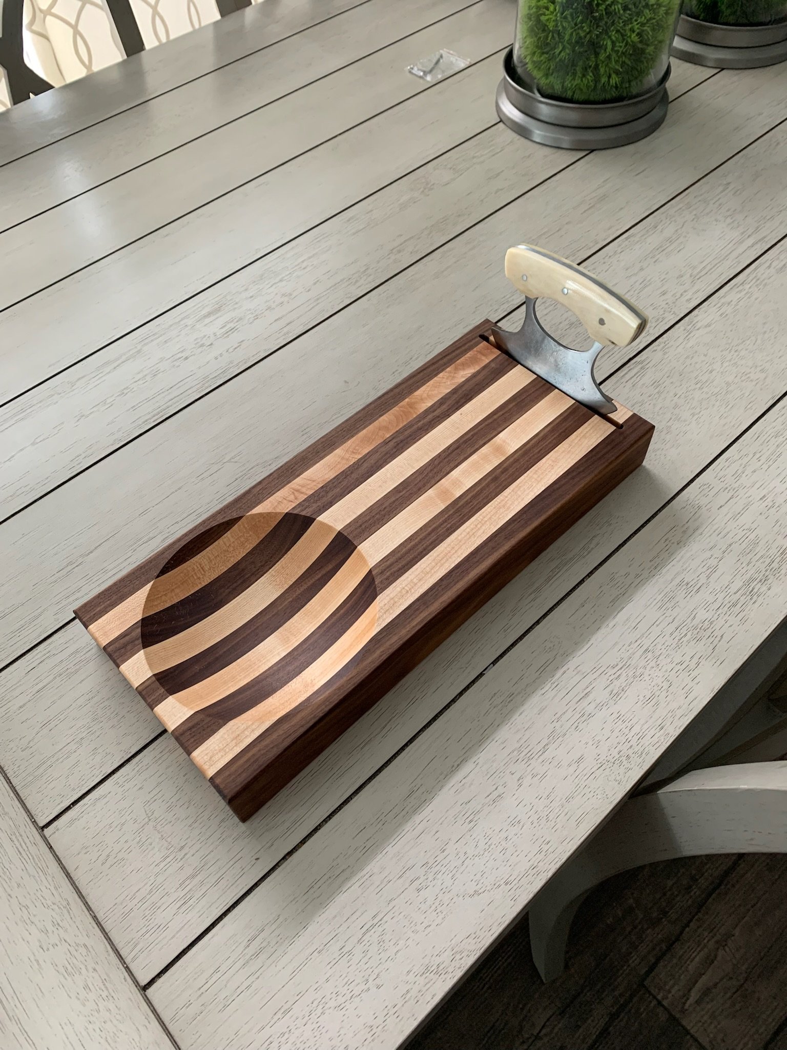 Wooden cutting board with handle on light-colored wooden dining table, with a glass container and potted plants in the background.