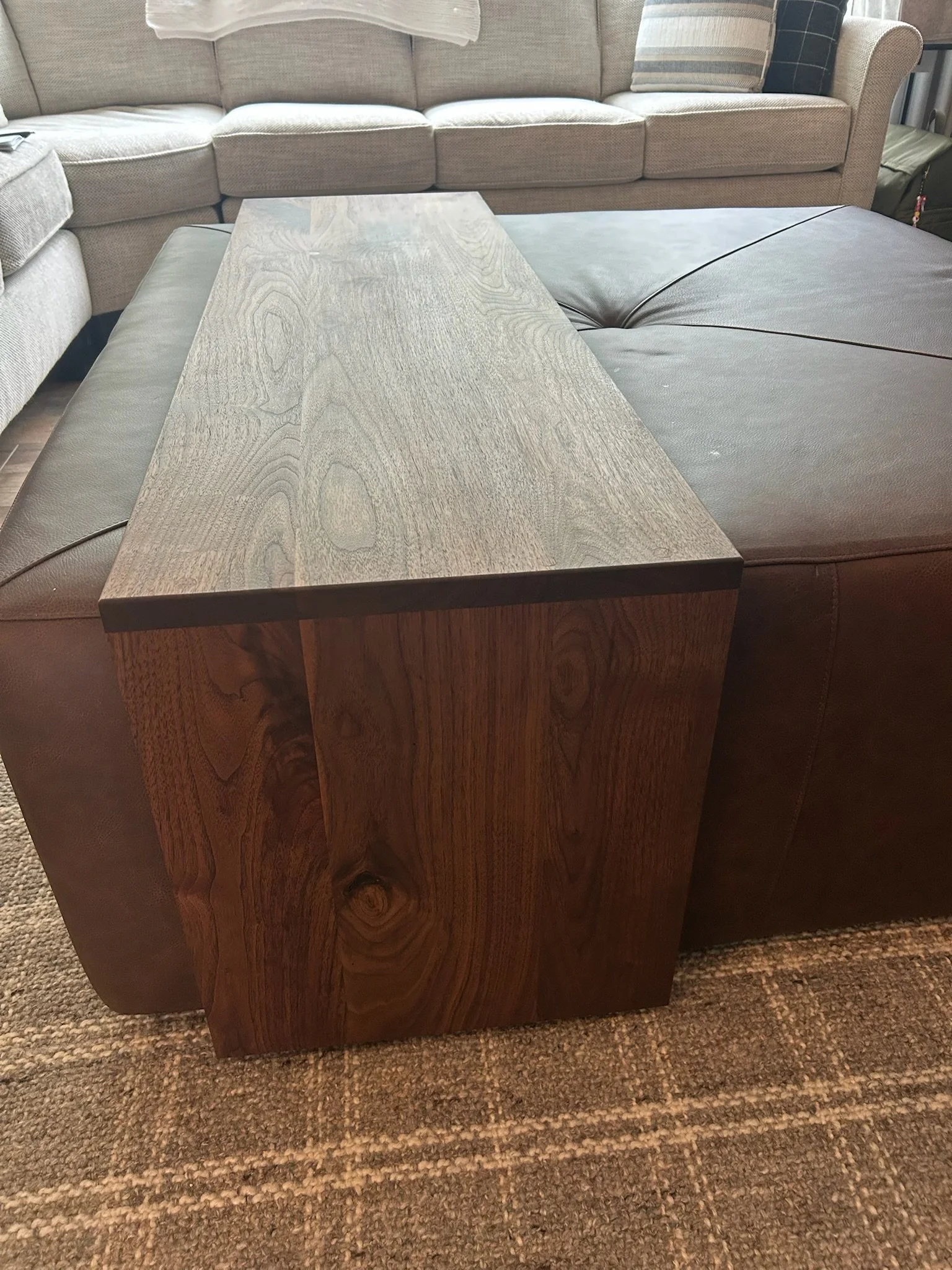 A rectangular wooden table with a visible wood grain, placed on a beige and brown patterned carpet. Part of a brown leather sofa is visible on the left, and a beige upholstered sectional sofa with pillows is in the background.
