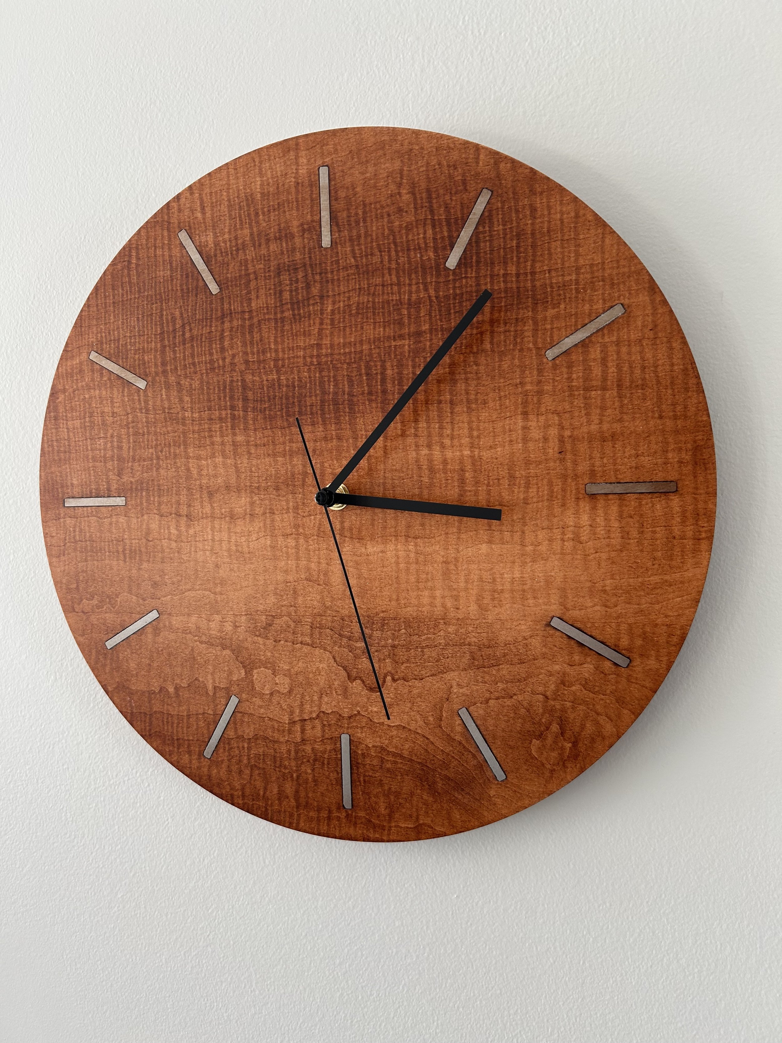 A round wooden wall clock with minimalist silver hour markers and black hour and minute hands, showing the time as approximately 3:07, mounted on a plain white wall.