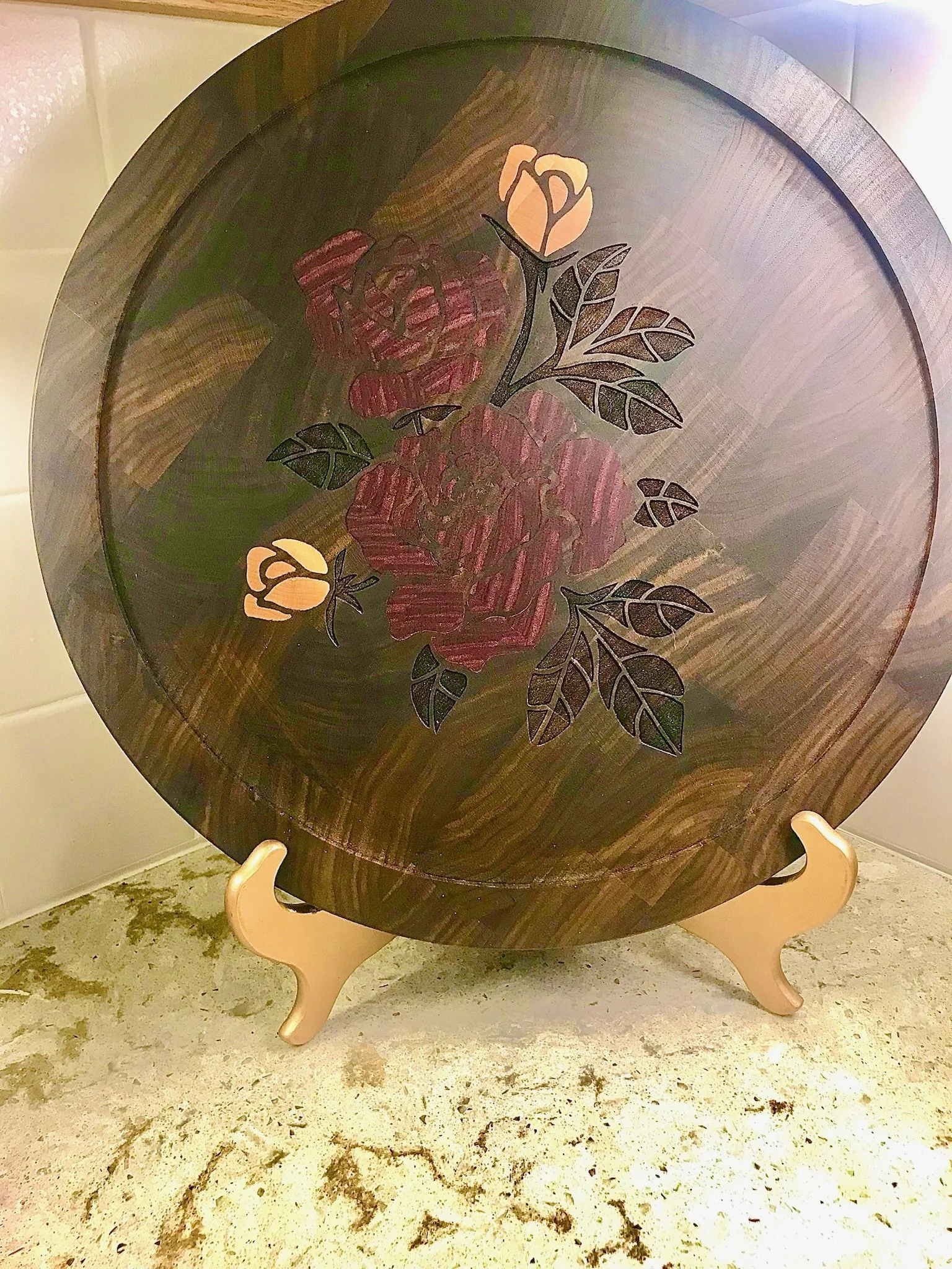 A round wooden table with a floral design featuring large pink roses and yellow buds, supported by three pendant-shaped light-colored wooden legs.