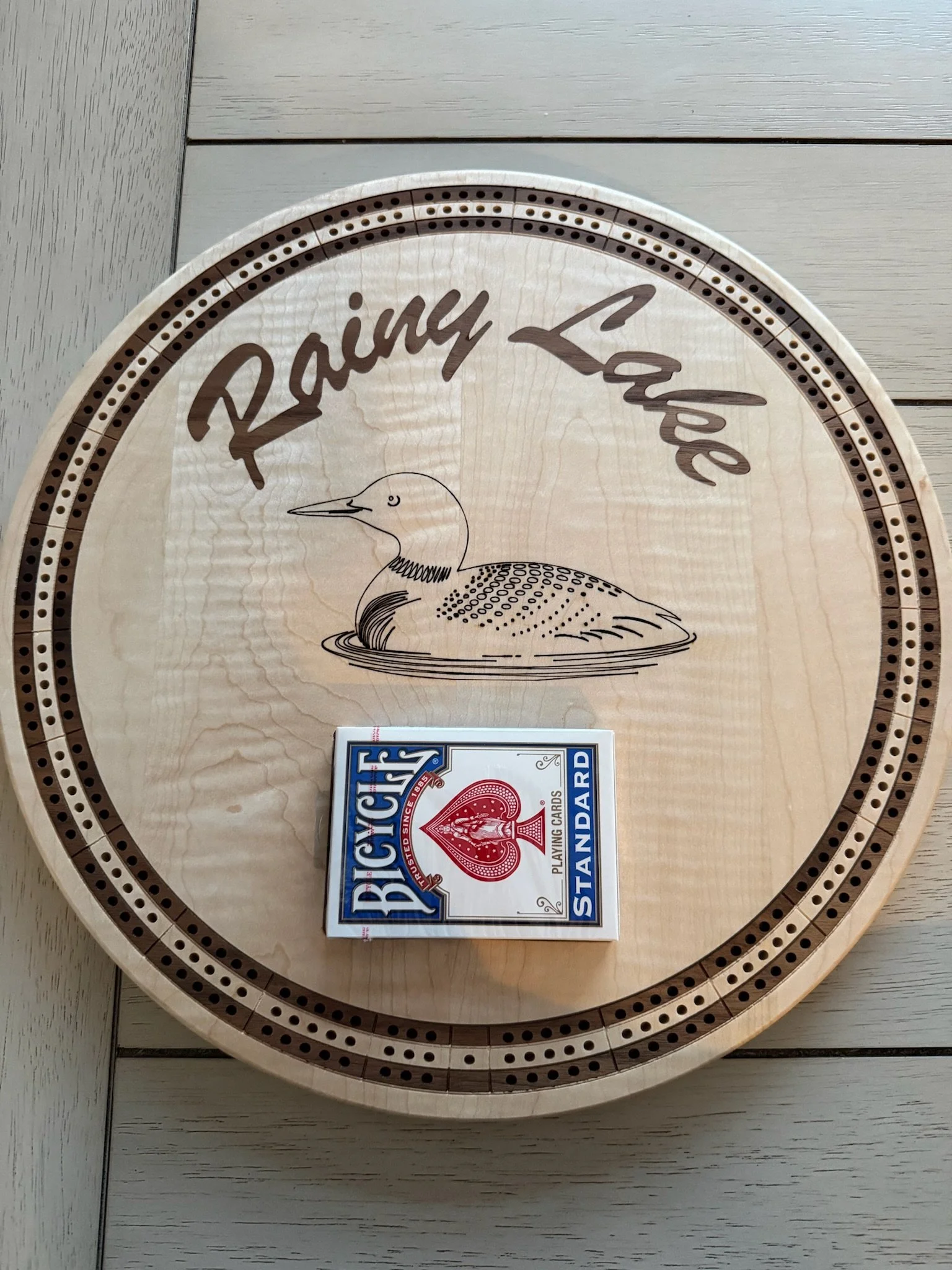 A circular wooden Raining Lake branded shuffleboard with a graphic of a loon swimming in water and a pack of Bicycle playing cards resting on it.