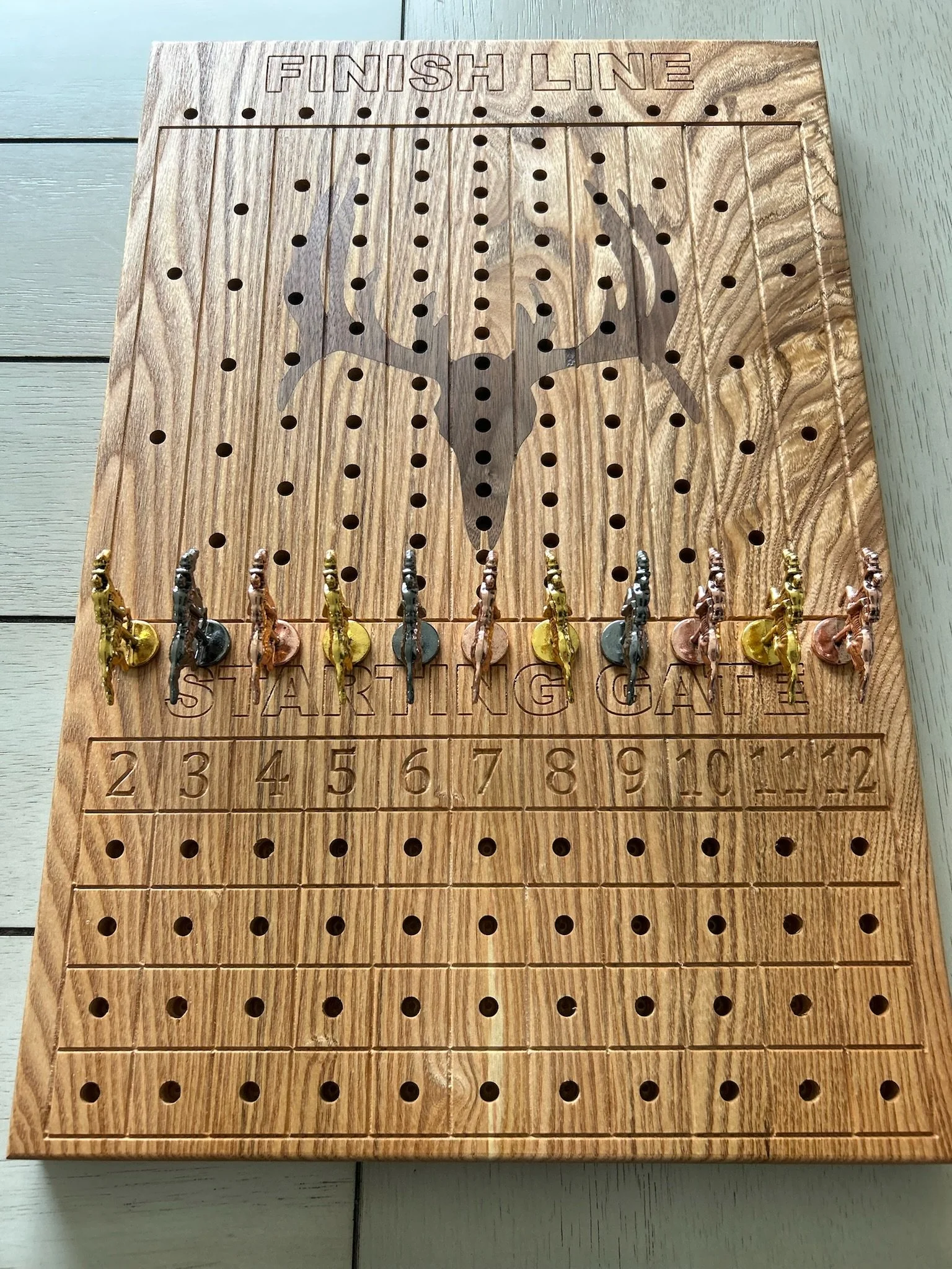 A wooden cornhole game board with holes, a deer skull design, and colored pegs at the bottom, with the words 'Finish Line' and numbered sections for scorekeeping.