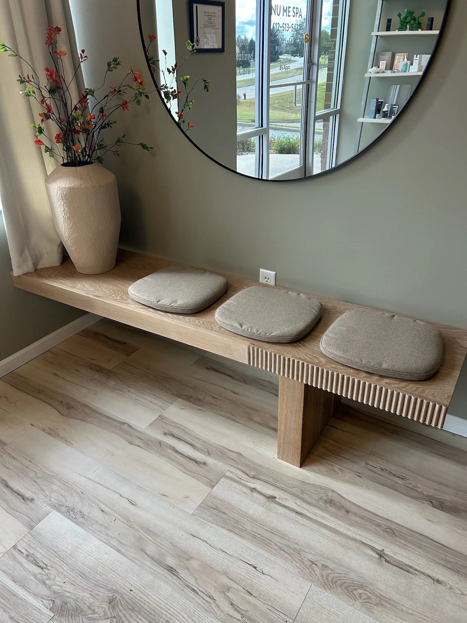 Interior view of a waiting area with a wooden bench with beige cushions, a large round mirror on a green wall, a tall beige vase with branches and red flowers, and a window reflecting the outside scene.