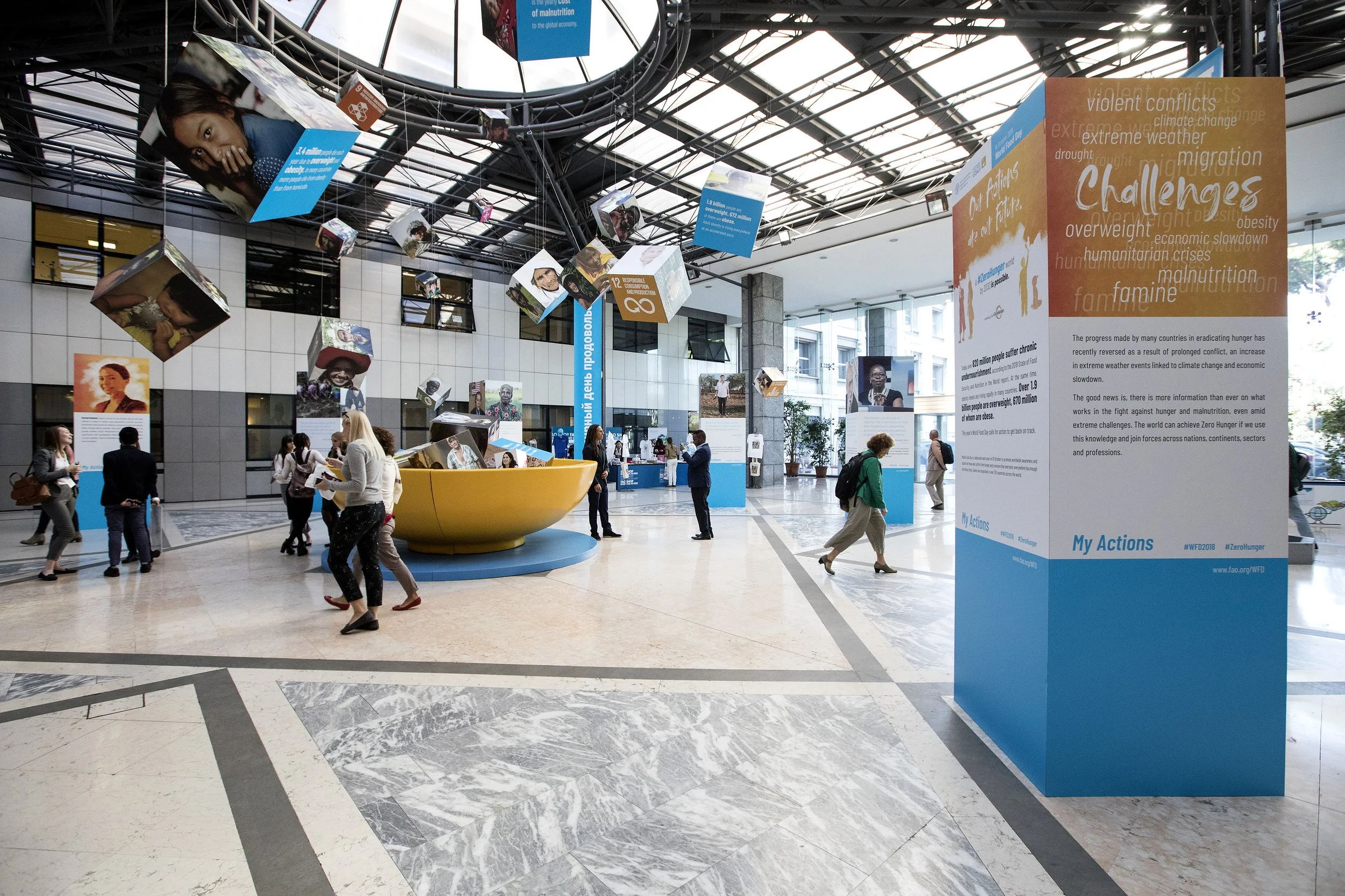 People gathering around an informational display at an indoor event about global issues such as climate change, migration, and hunger, with hanging posters and photos from the ceiling.