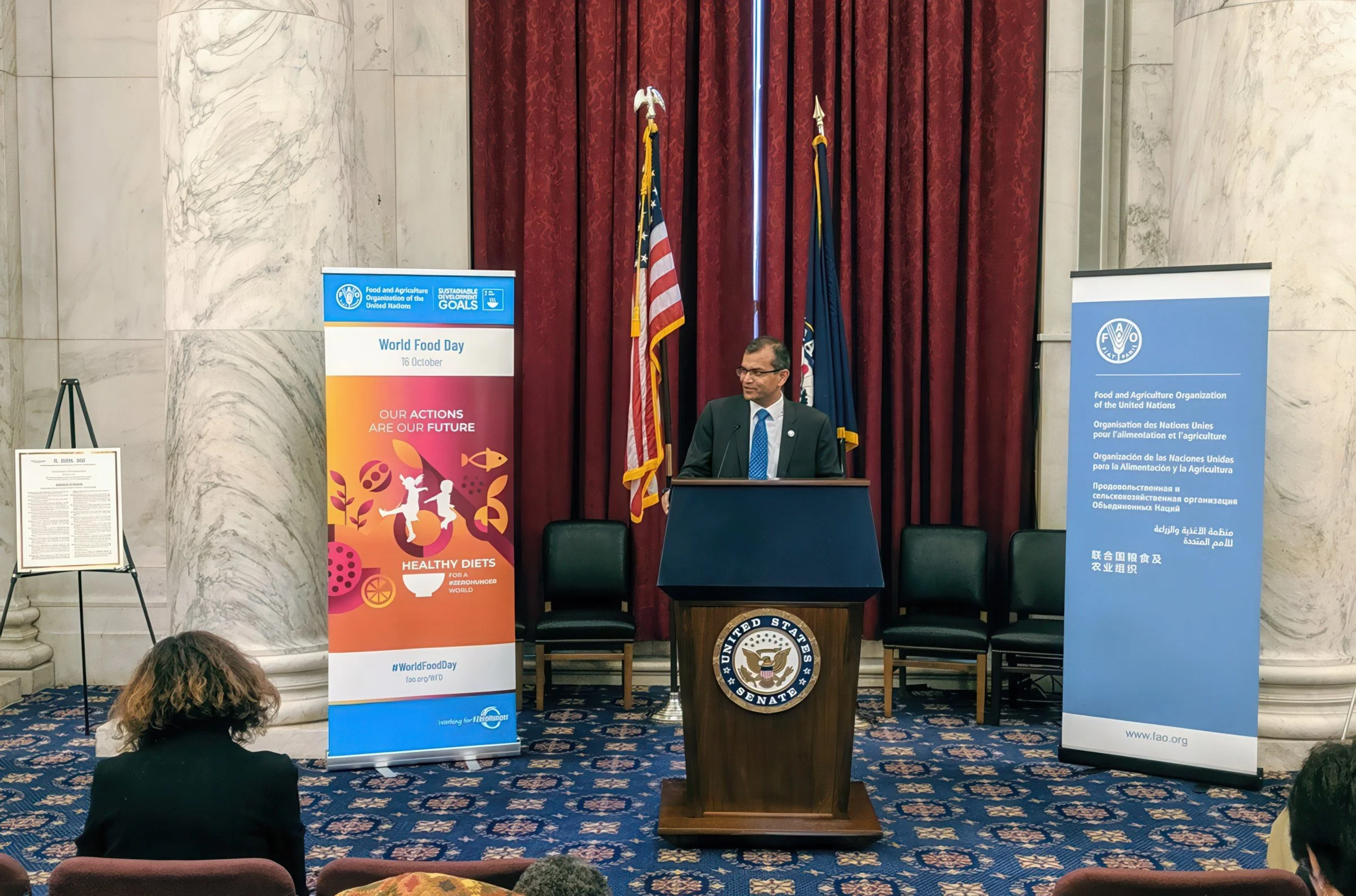 A man standing at a podium with the United States Senate seal, giving a speech or presentation. Behind him are two flags and maroon curtains. To the left and right of him are banners promoting World Food Day and the Food and Agriculture Organization 
