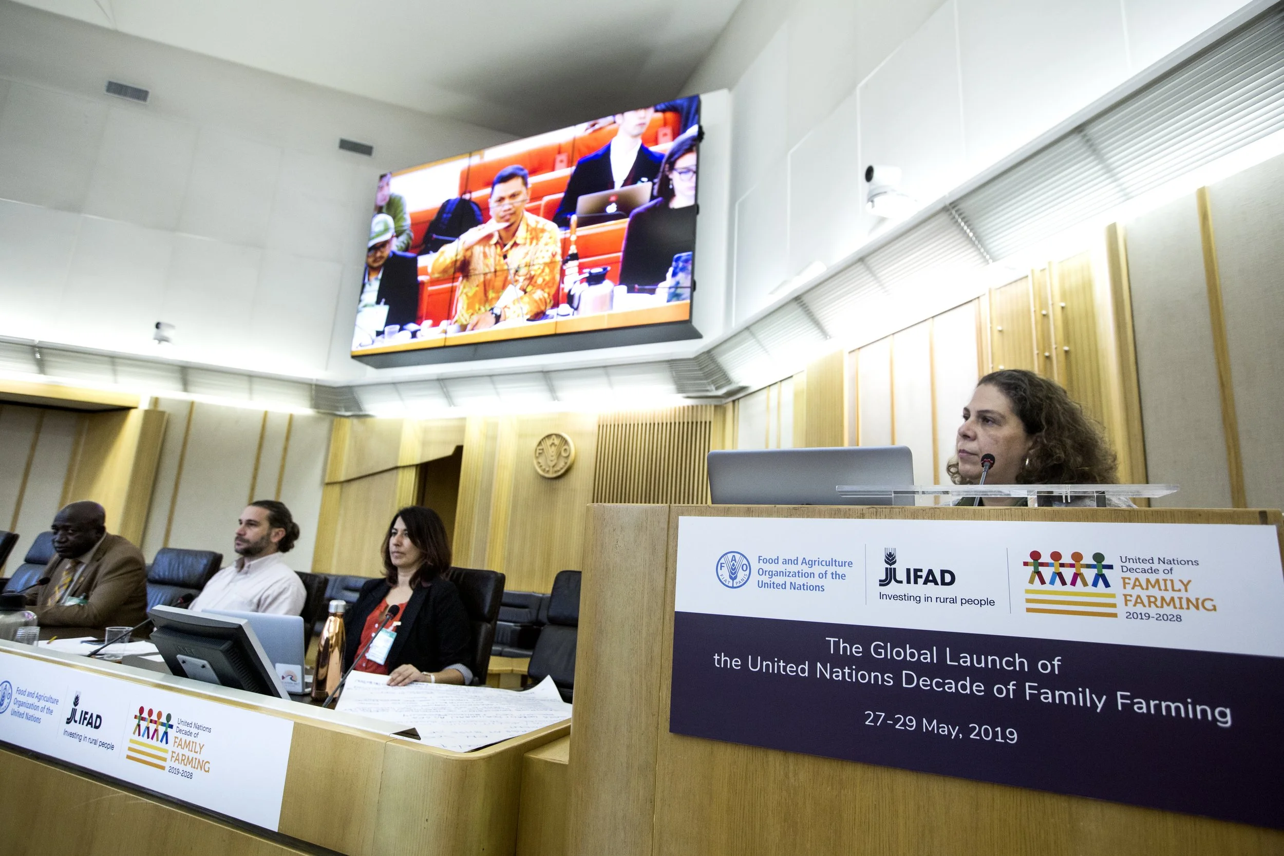 Conference room with panel of five people sitting at a long table, woman with curly hair speaking at a podium with banners showing logos of UN FAO, UN Decade of Family Farming, and IFAD; large screen behind displaying a zoom call with more participan