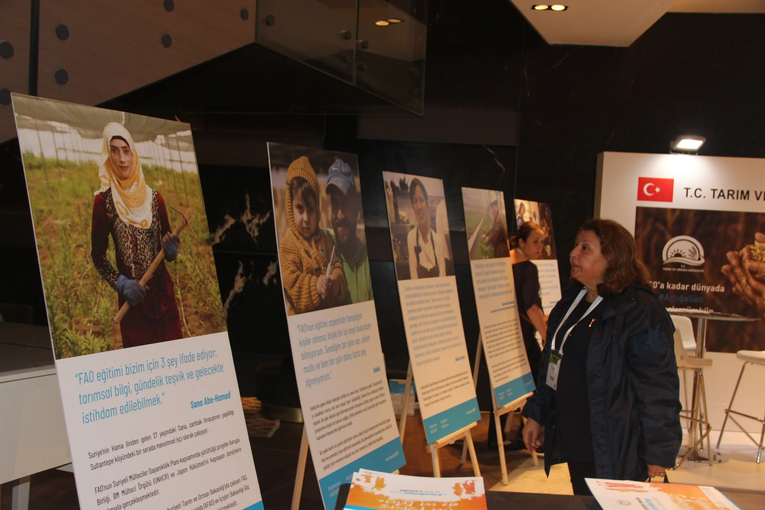 An indoor exhibition with display boards featuring photographs and information. Several people are viewing the displays, one woman in dark clothing and a lanyard is in the foreground, engaging with a display. The setting appears to be a conference or
