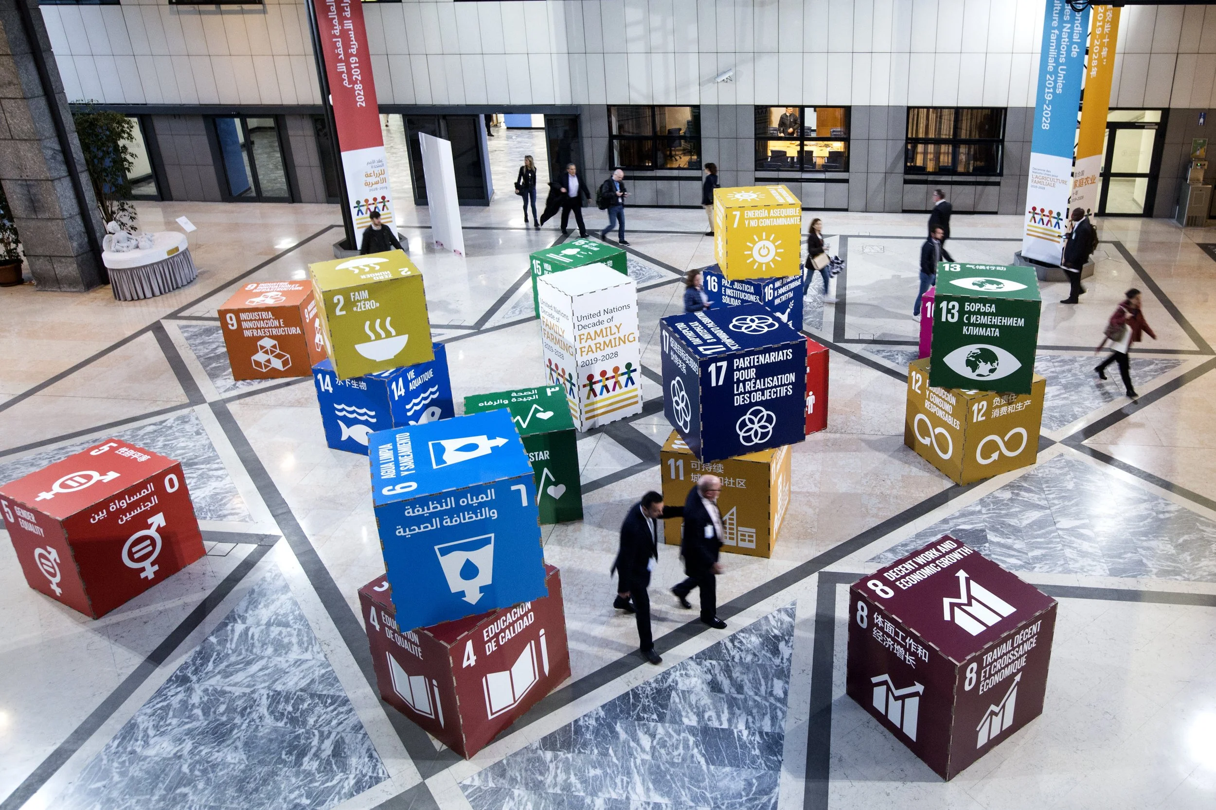 An indoor lobby area with large colorful cubes displaying sustainable development goals. People are walking and talking, and some are looking at the cubes.