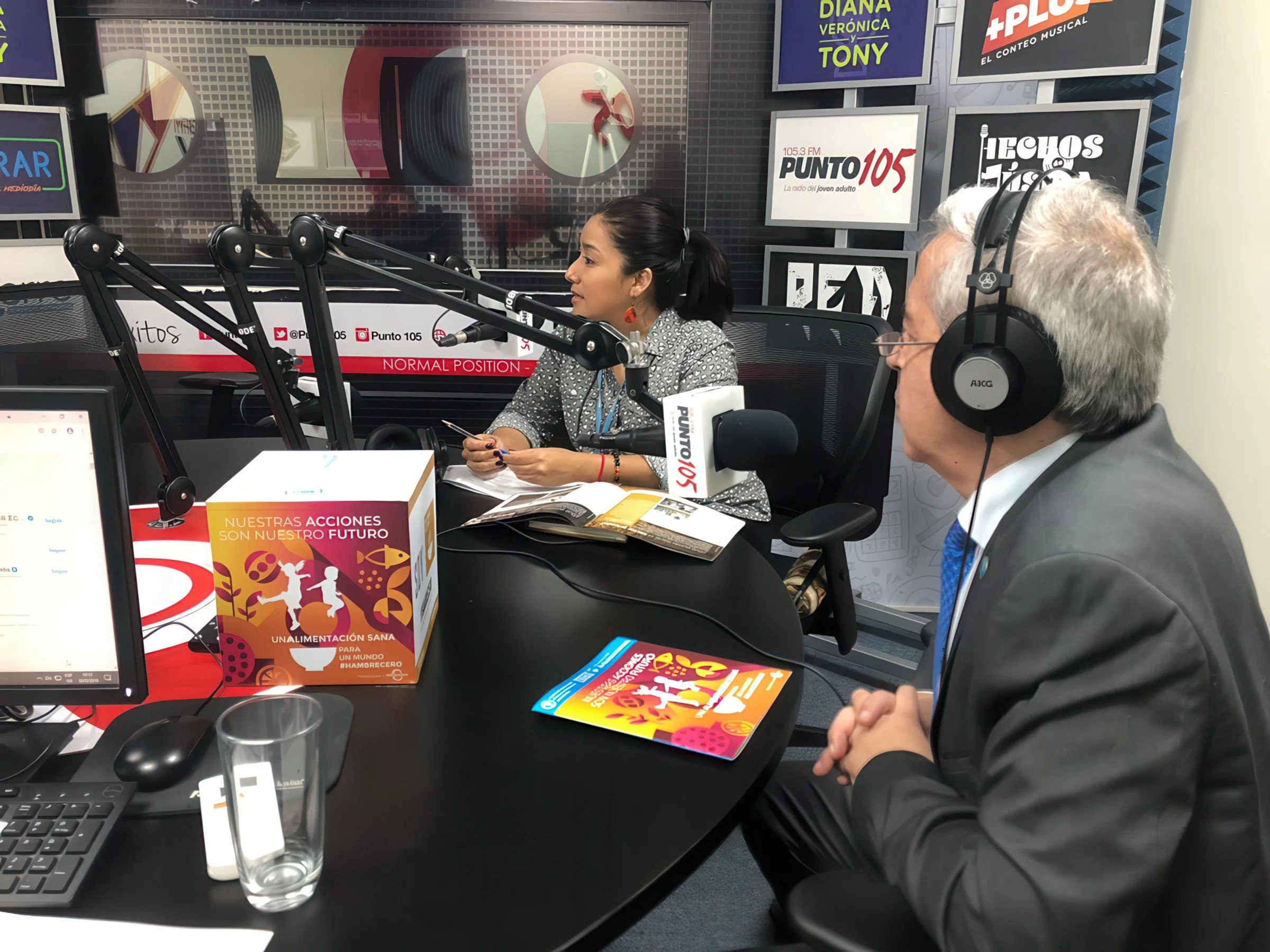 Radio station studio with two people speaking into microphones, one woman with dark hair and the man with gray hair and glasses, surrounded by posters on the wall promoting radio shows and campaigns, with recording equipment and a glass of water on t