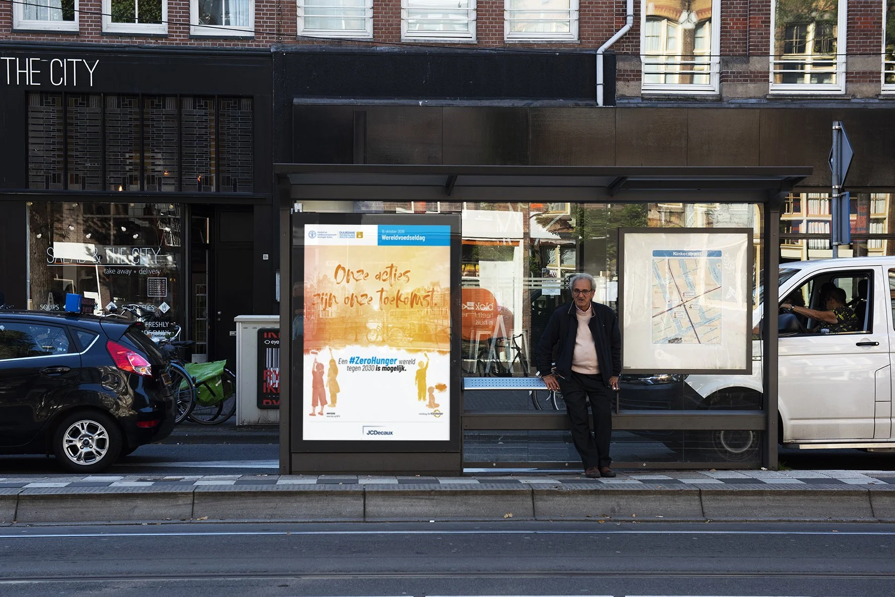 A city bus stop with an advertisement poster and a map on the side. There is a man standing at the bus stop, and a white van and a black car are visible behind the bus stop. A man is sitting inside the white van, and a woman is sitting on a bicycle b