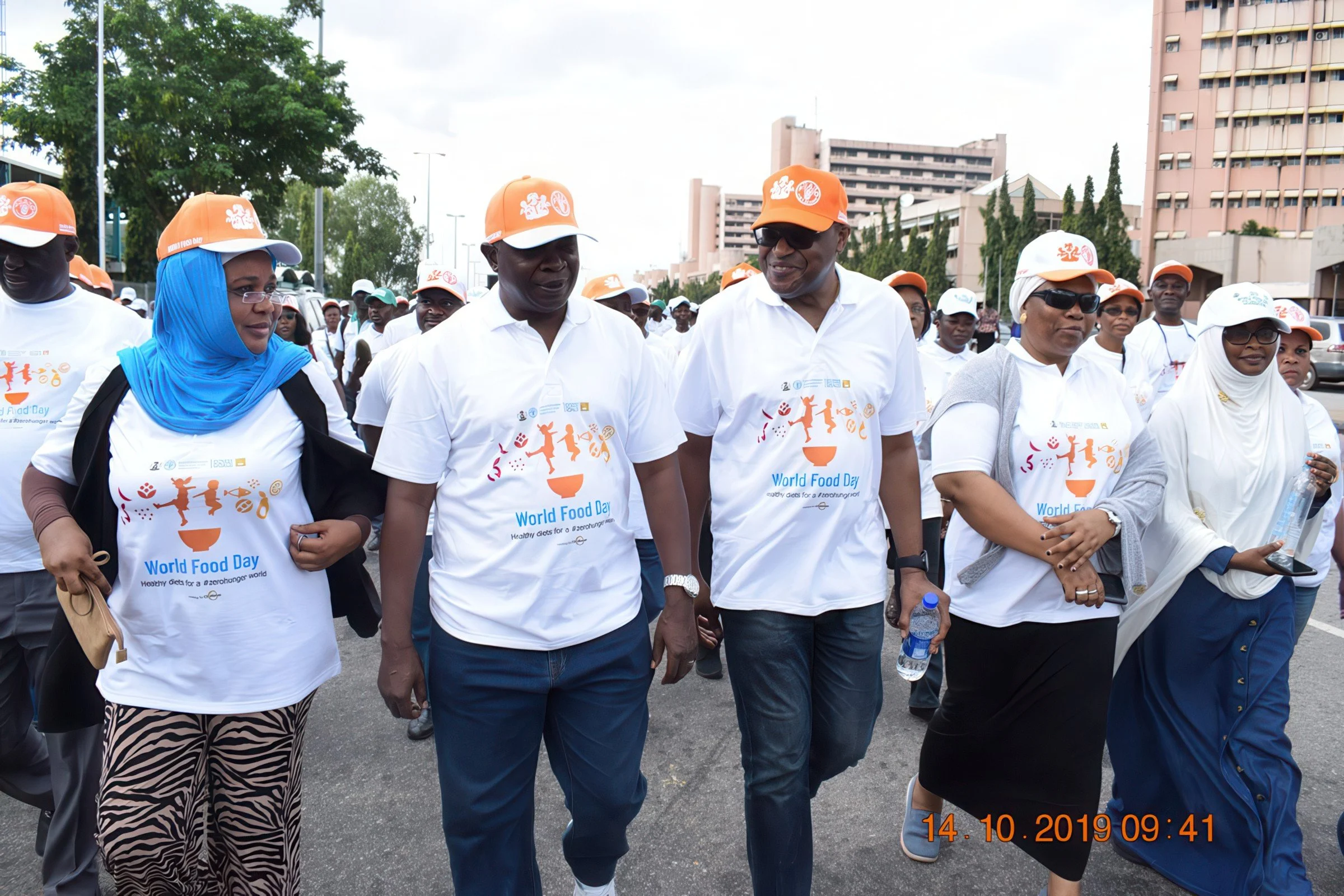 A group of diverse people participating in a World Food Day event, walking outdoors in an urban area, wearing white T-shirts with the event logo and orange caps.