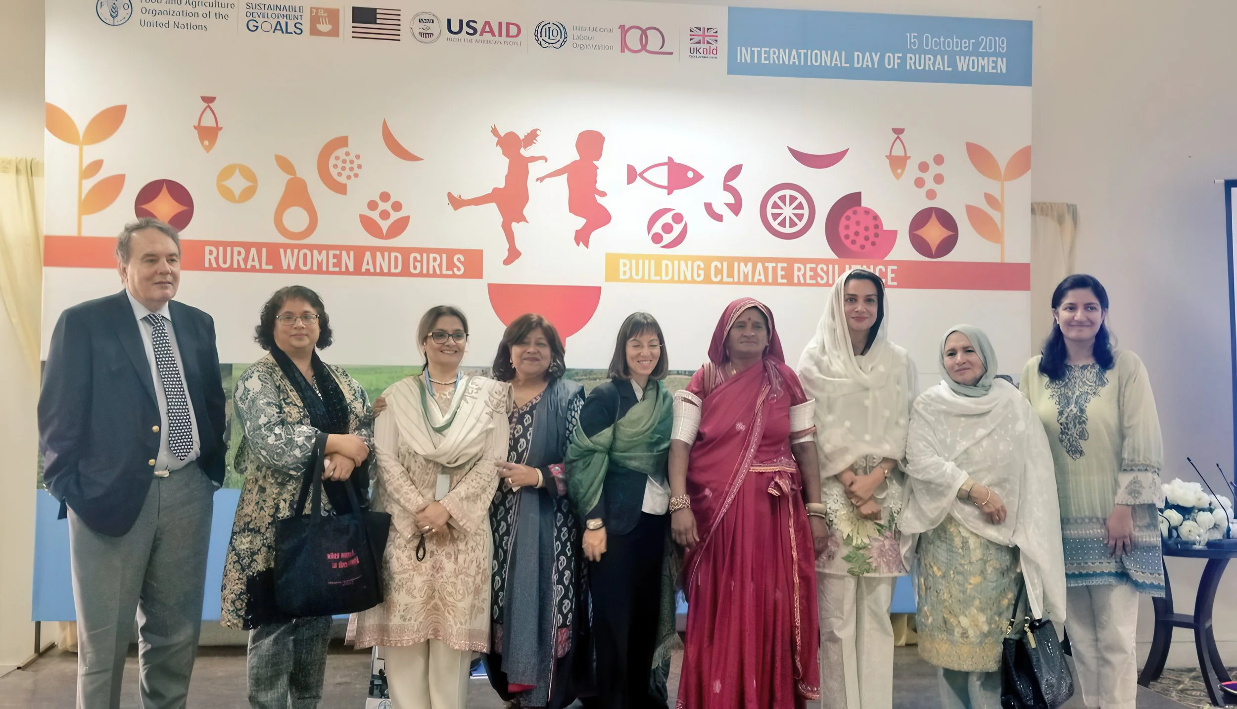 A group of diverse women and a man standing together in front of a colorful banner that reads, "International Day of Rural Women, 15 October 2019," with various logos and symbols, including images of children, flowers, and fish.
