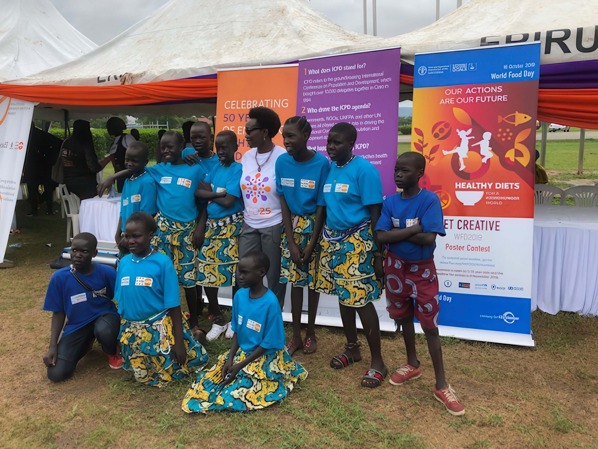 Group of children and a woman standing in front of an informational booth at an outdoor event, with colorful banners promoting healthy diets and celebrating 50 years of an organization, on a grassy area under a tent.