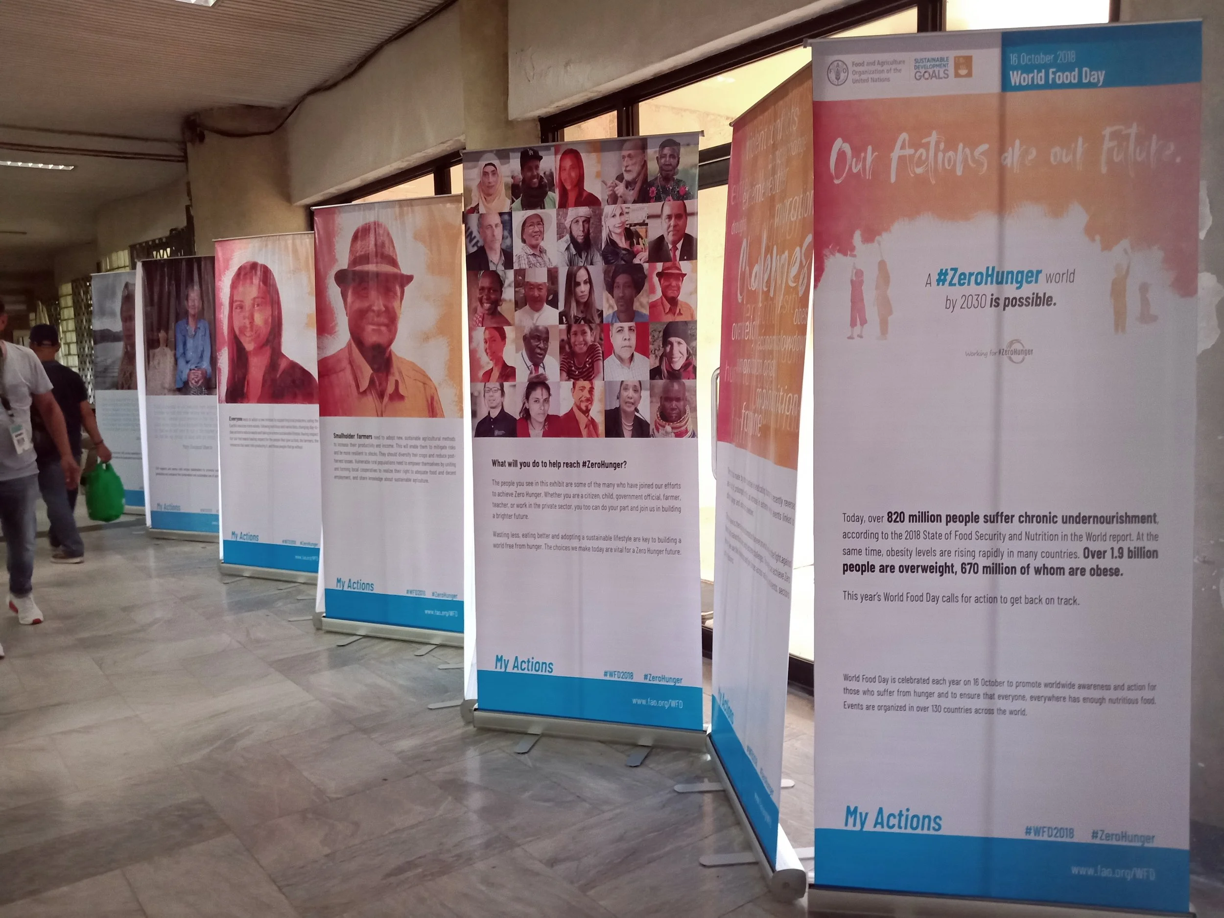 Banners displayed at an indoor event showcasing portraits of diverse individuals and information about World Food Day, sustainable development goals, and zero hunger initiative with people walking in the background.