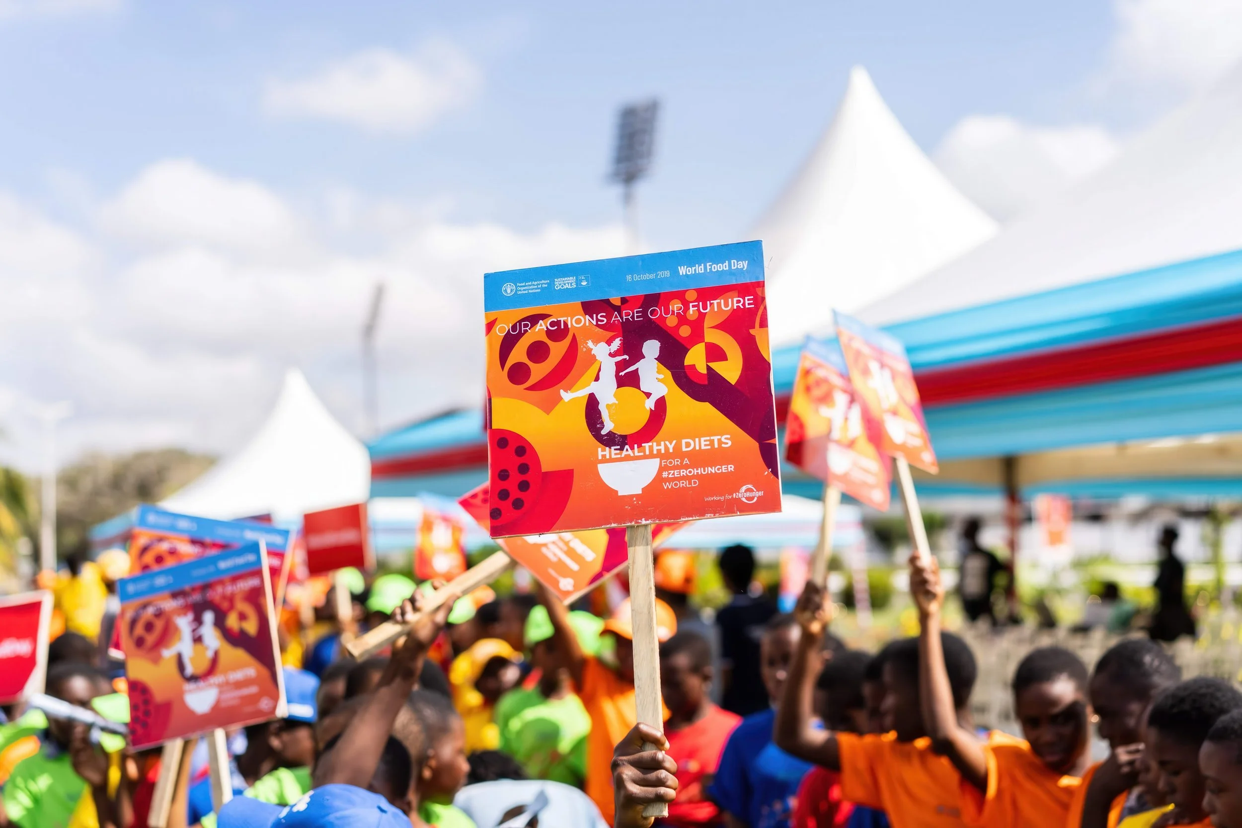 A crowd of people holding World Food Day banners at an outdoor event with tents and a partly cloudy sky.