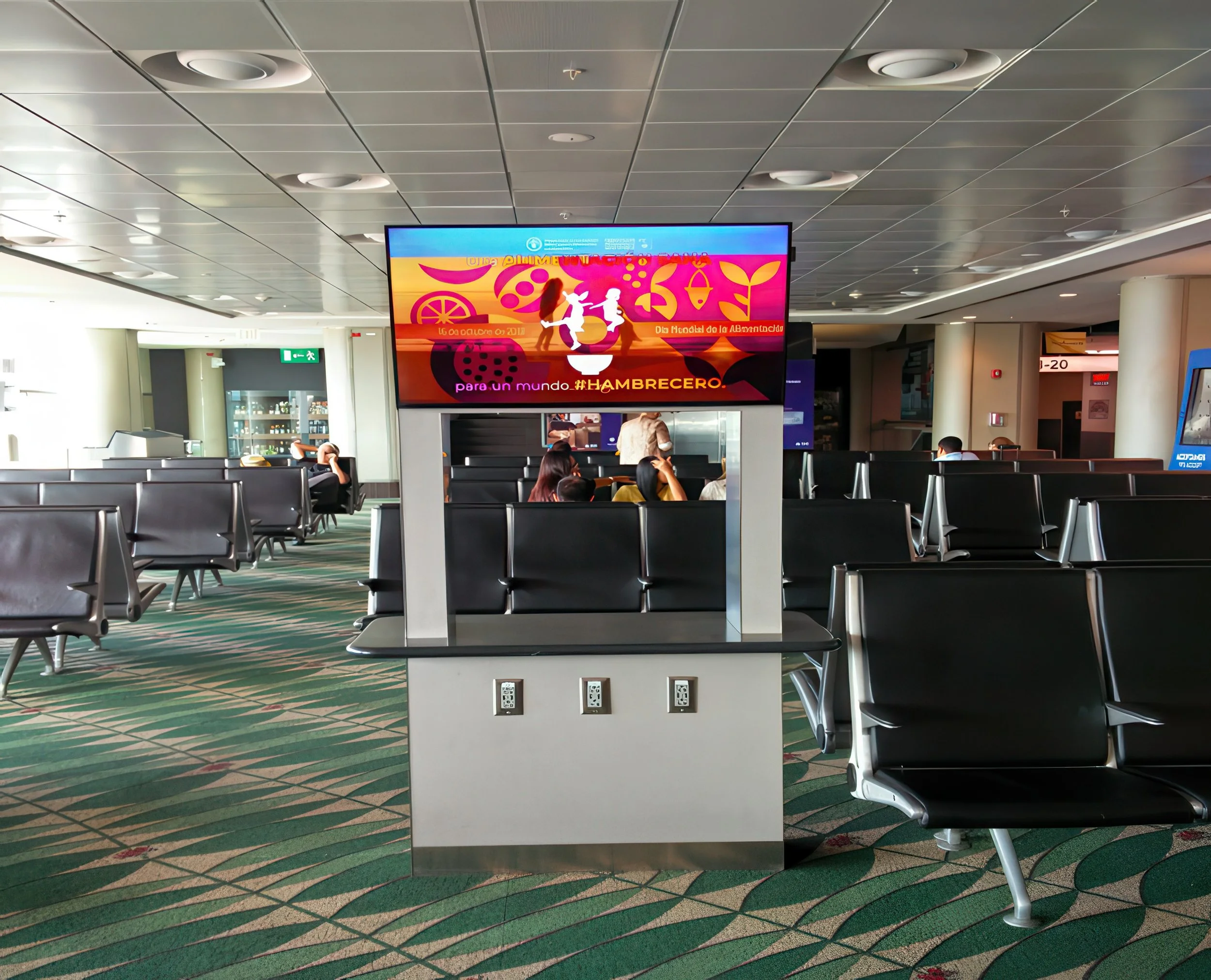 An airport waiting area with black chairs, a TV screen displaying colorful graphics, and a few people sitting and waiting. The ceiling is gray with circular light fixtures, and there is a vending machine and an exit sign in the background.